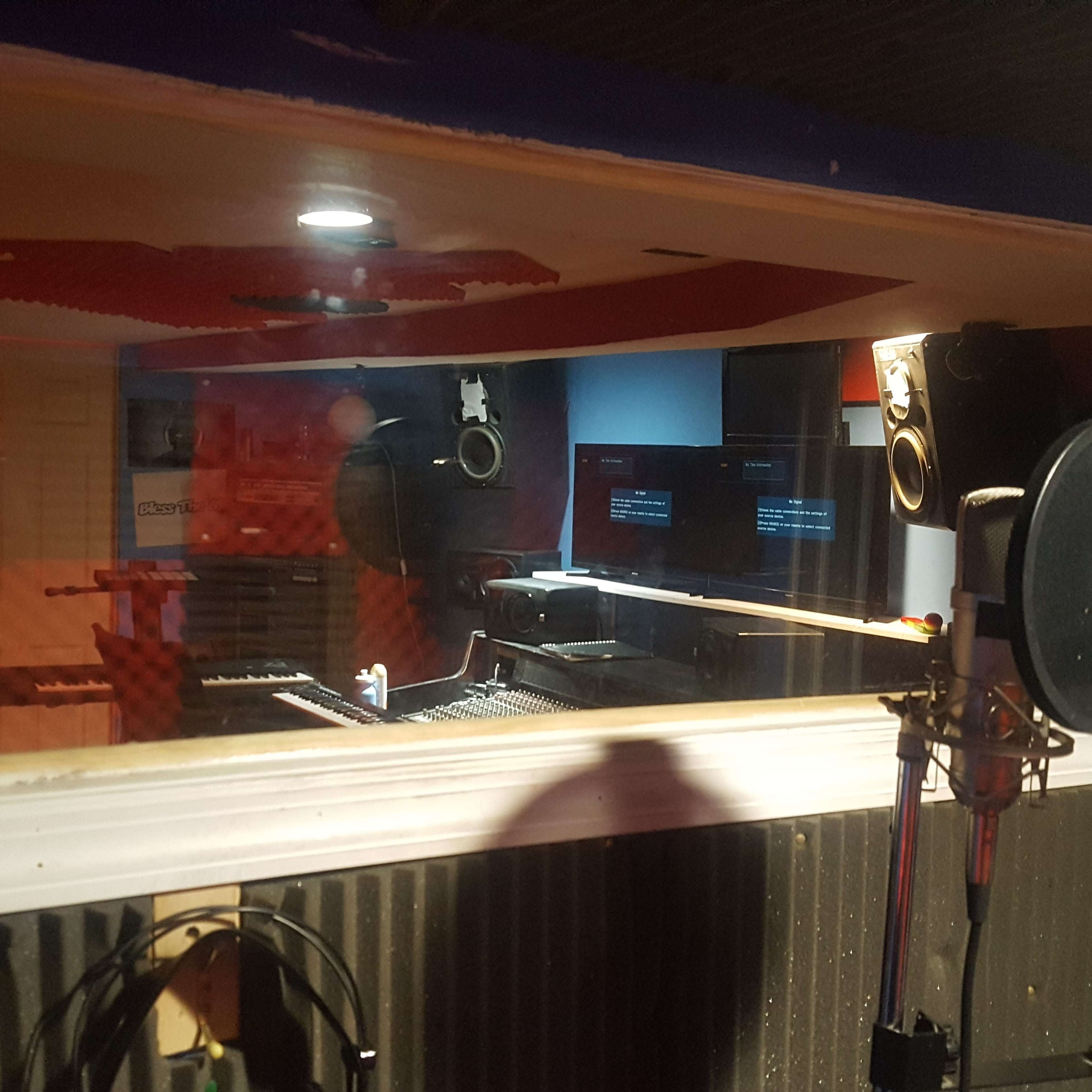 A recording studio with a control desk, monitors, and microphones visible through a glass window.