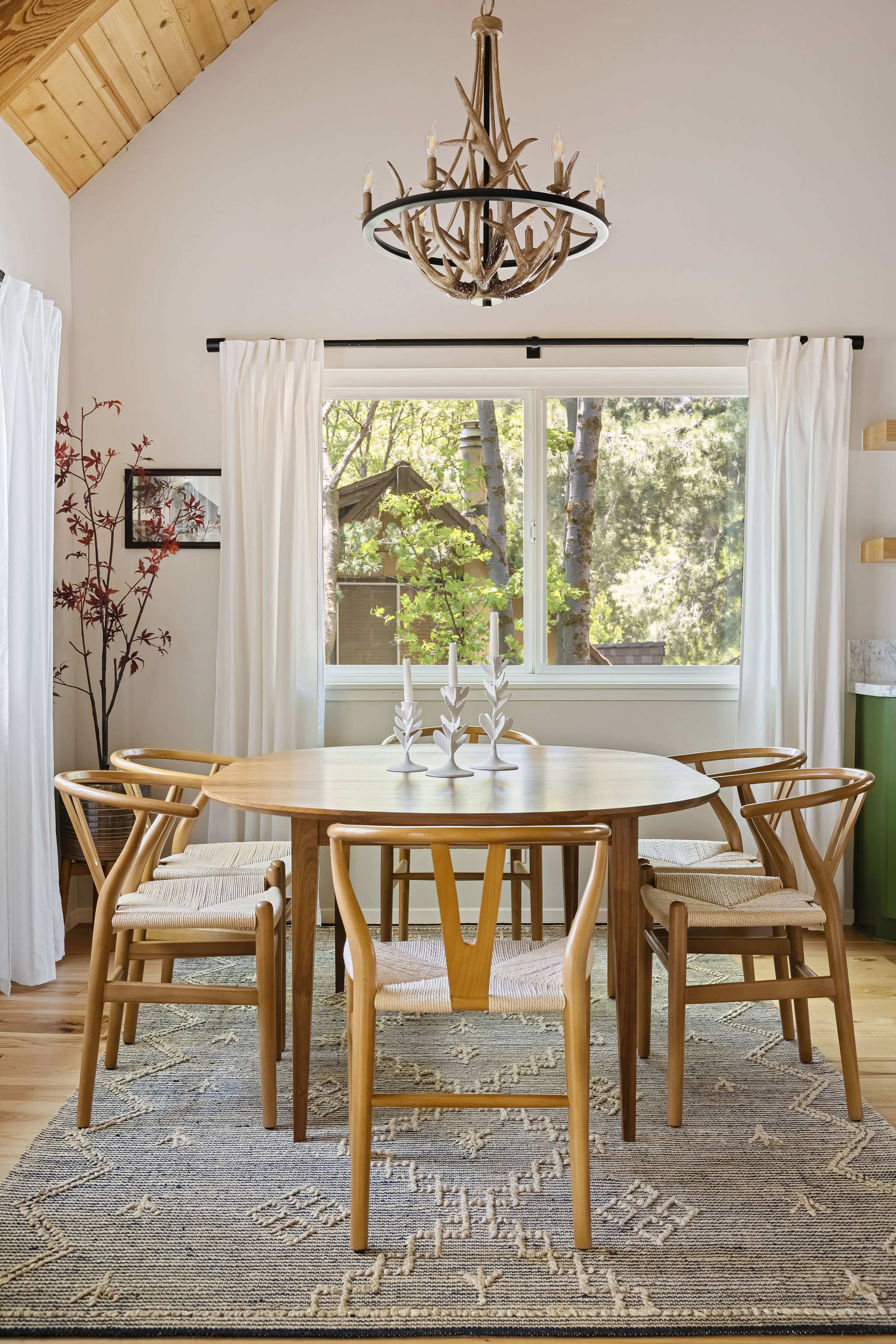 A round wooden dining table with four chairs is set up in a well-lit room featuring a large window and a chandelier overhead.