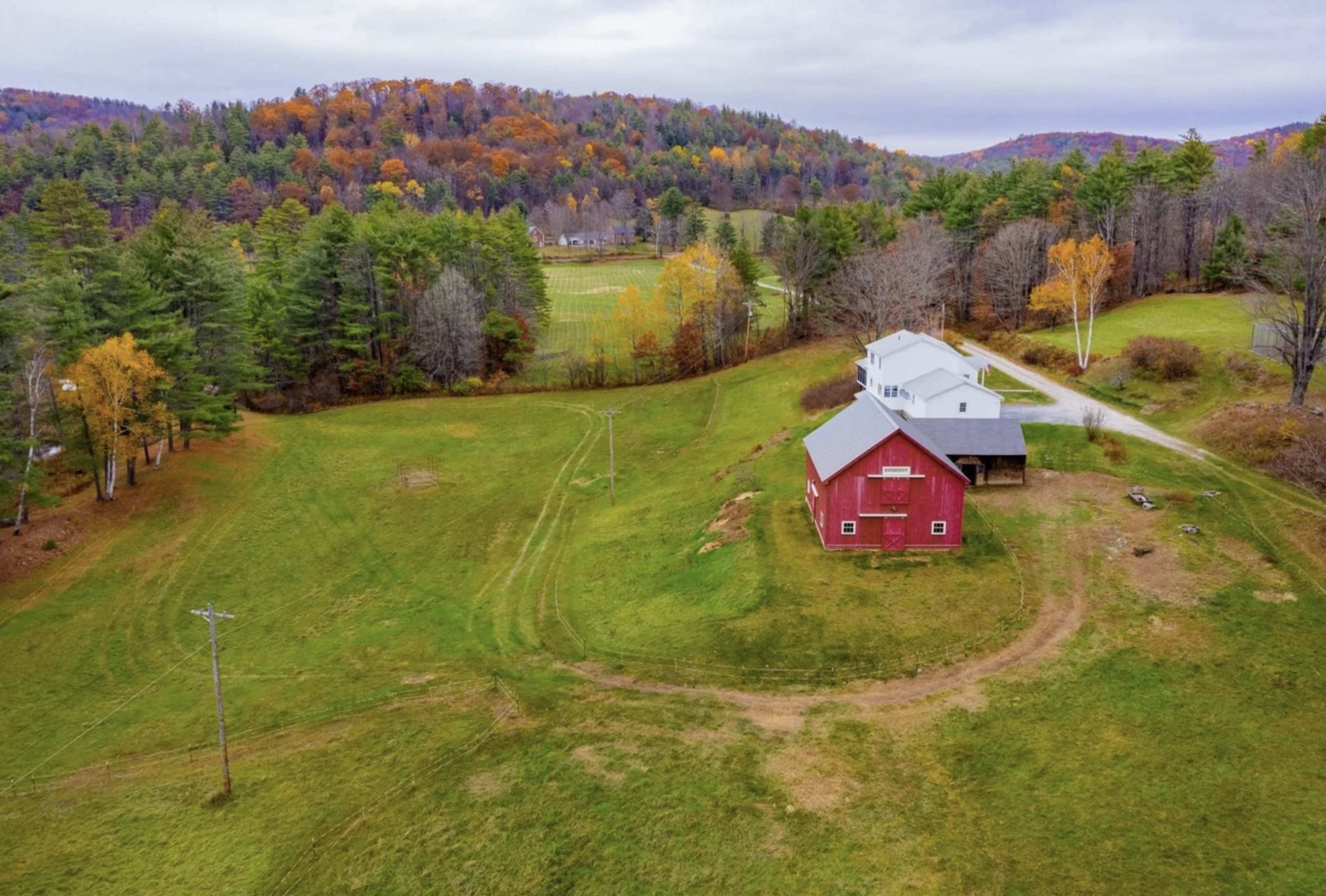 A red barn and a neighboring white house are situated on a grassy landscape surrounded by trees with autumn foliage.