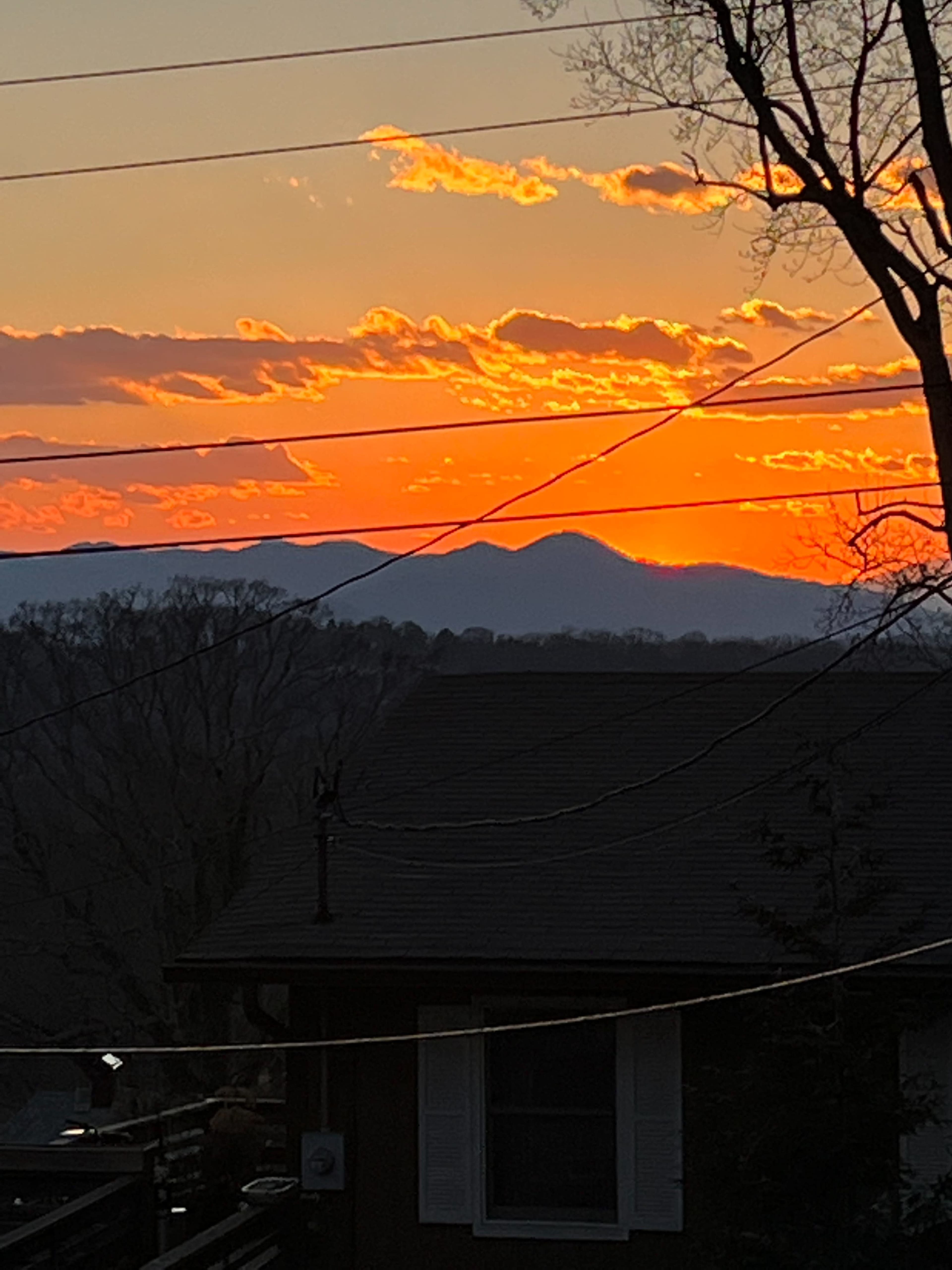 A sunset with vibrant orange and purple hues above a mountain range, framed by silhouetted trees and houses.