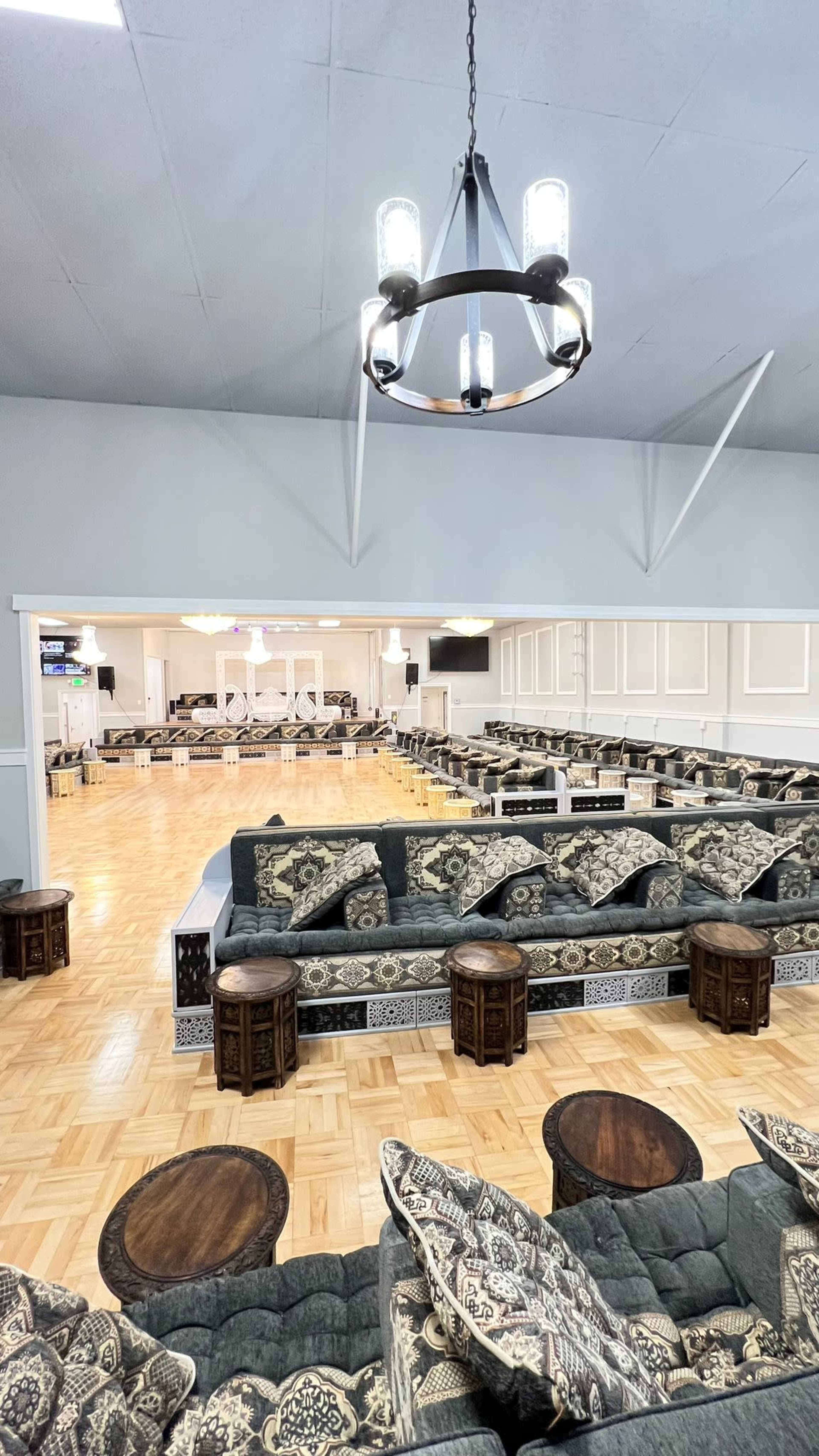 The image shows a spacious event hall with patterned seating arrangements, wooden tables, and a polished wooden floor.