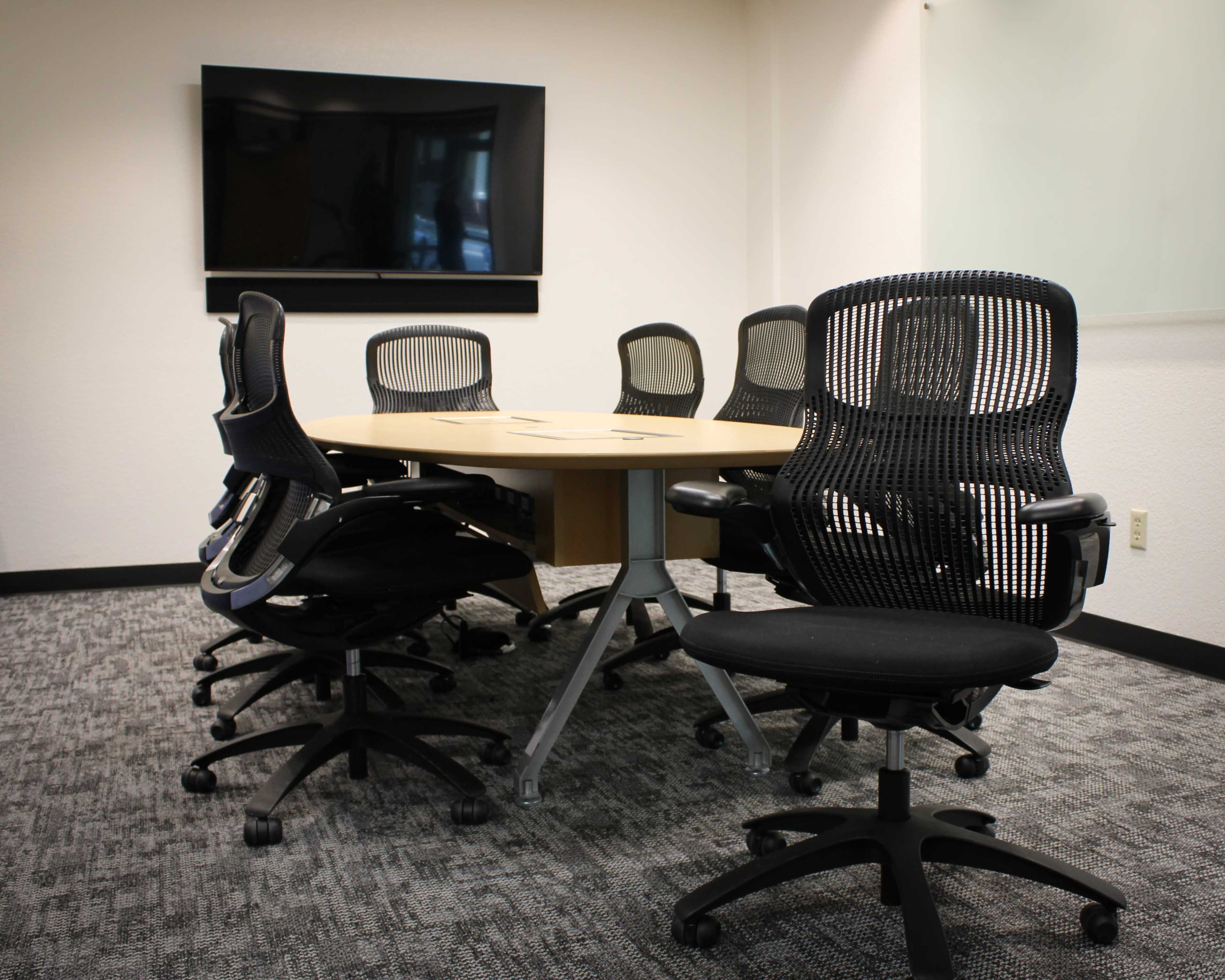 A circular conference table surrounded by black ergonomic chairs is situated in a well-lit meeting room with a television screen mounted on the wall.