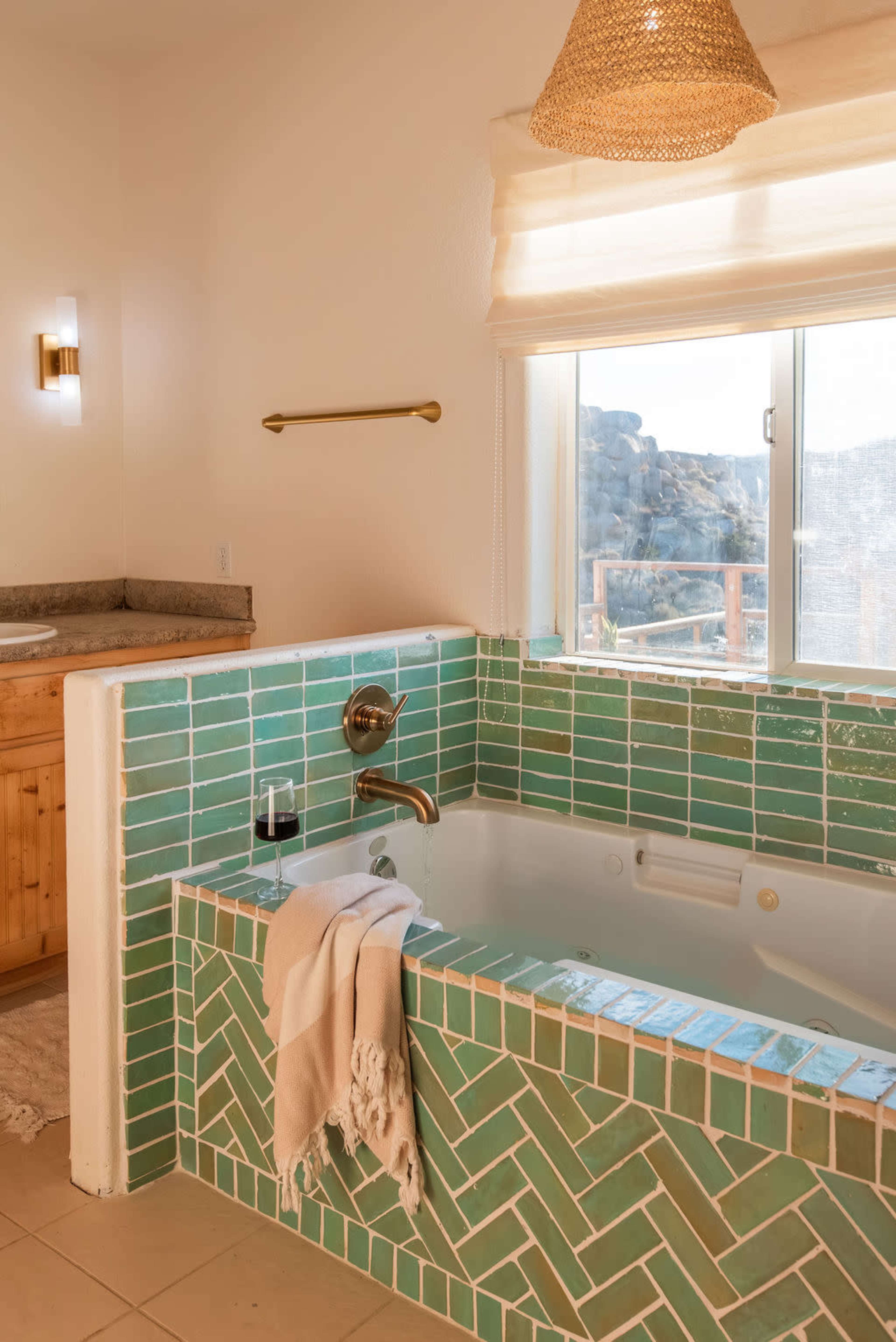 The image shows a bathroom featuring a green tiled bathtub with a beige blanket draped over it, a wooden vanity in the background, and a large window that lets in natural light and offers a view of rocks outside.