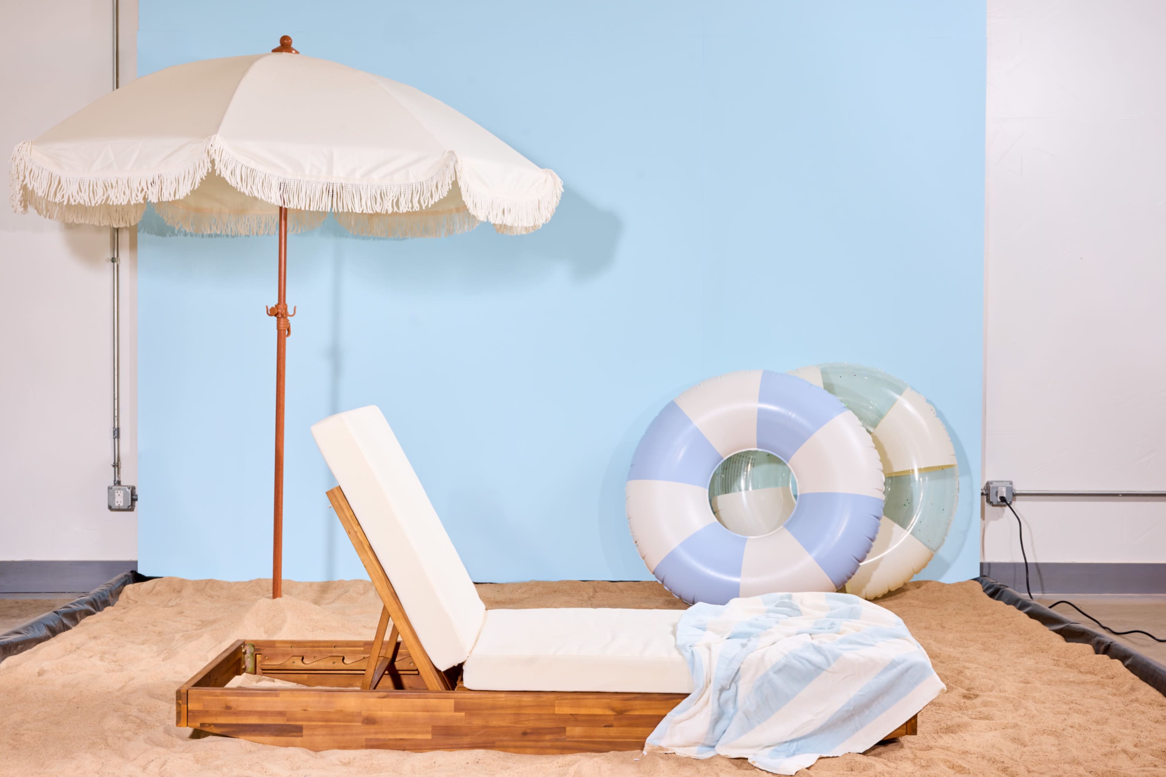 A beach scene features a wooden lounge chair, a large white umbrella, and two inflatable swim rings, all set against a light blue backdrop with sand underfoot.