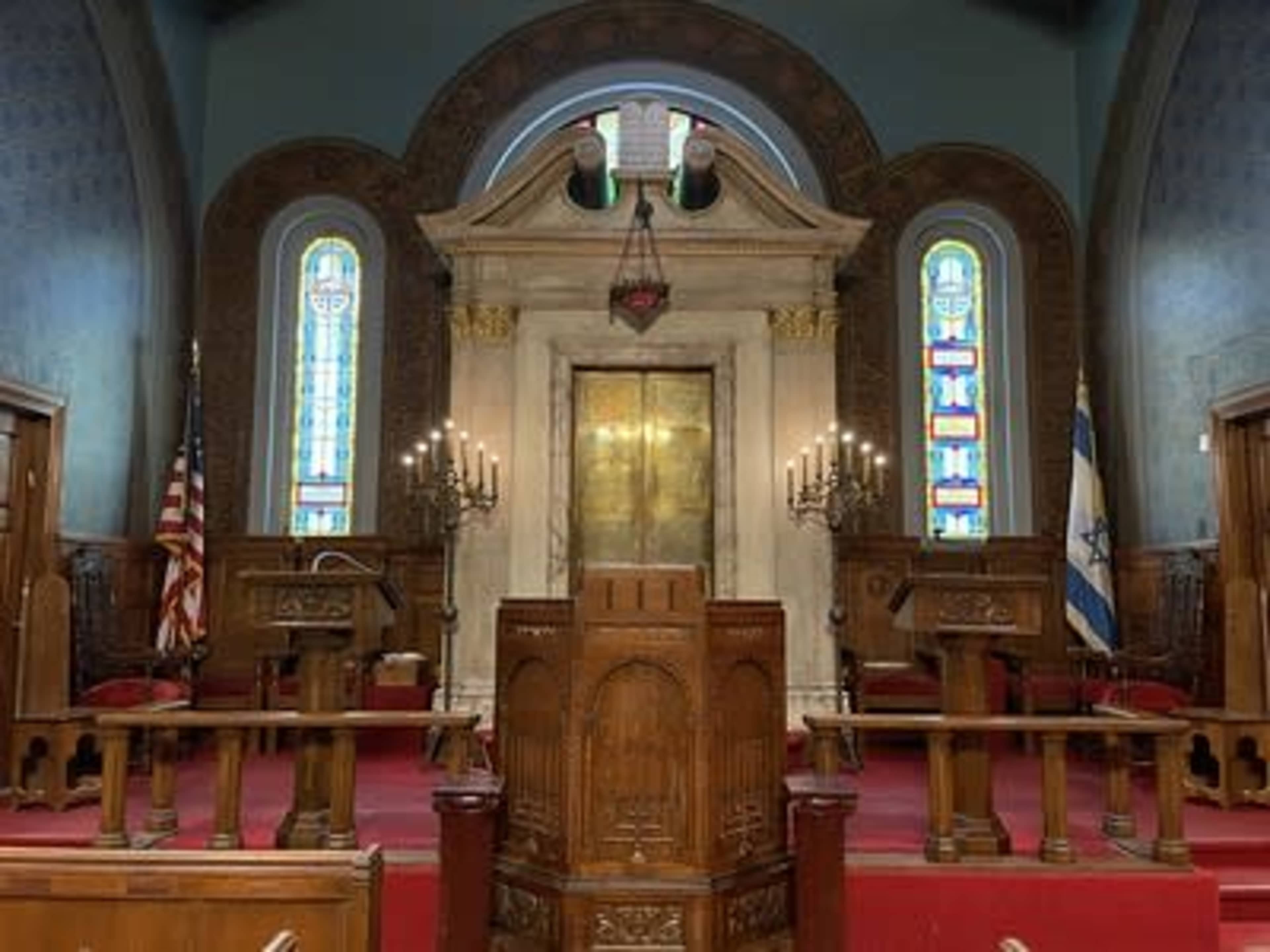 The interior of a synagogue features a pulpit in the foreground, flanked by wooden chairs, with ornate stained glass windows and a large central ark behind it.