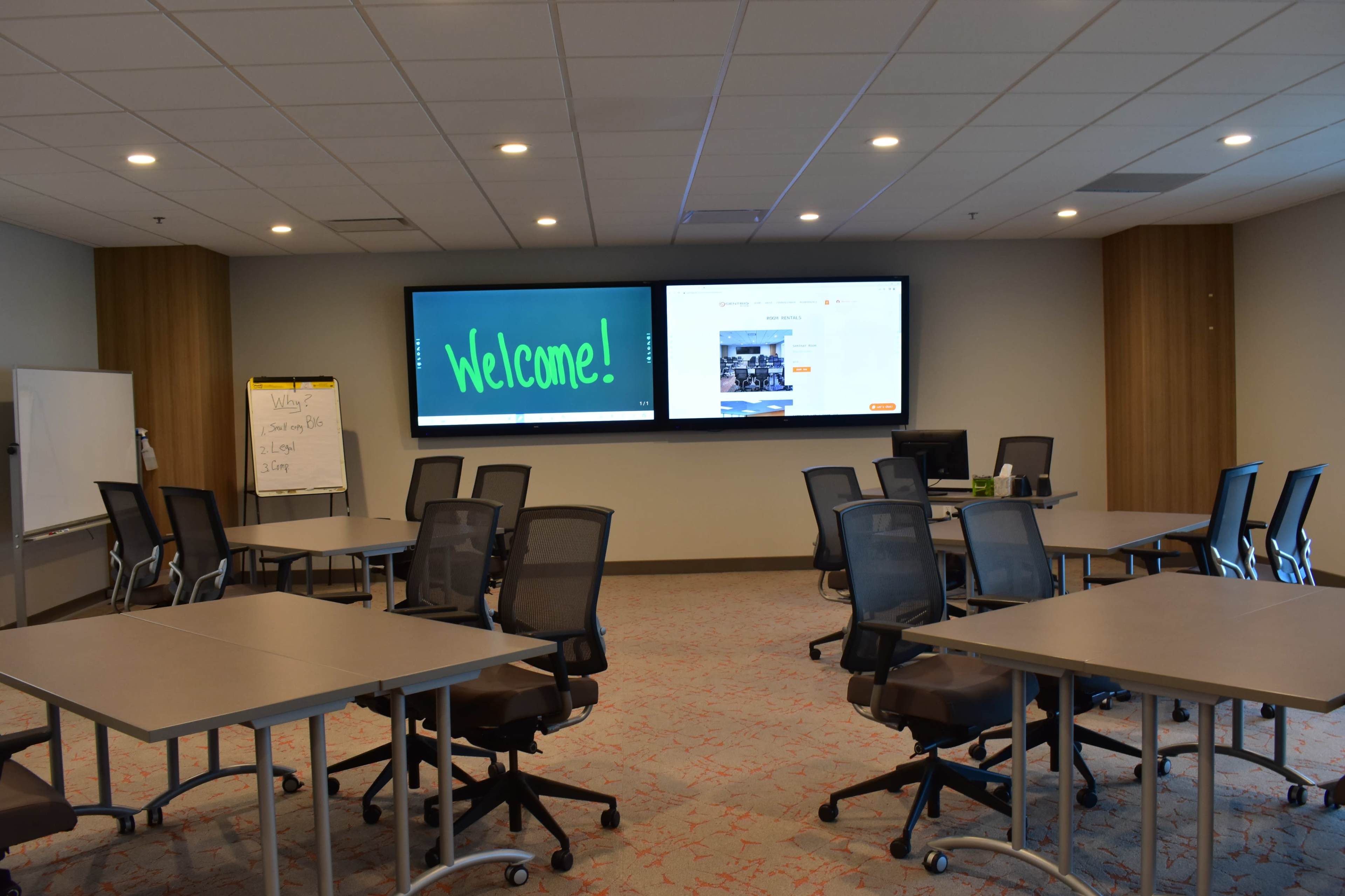 A modern conference room with tables arranged for a meeting, featuring two large screens displaying a welcome message and additional content.