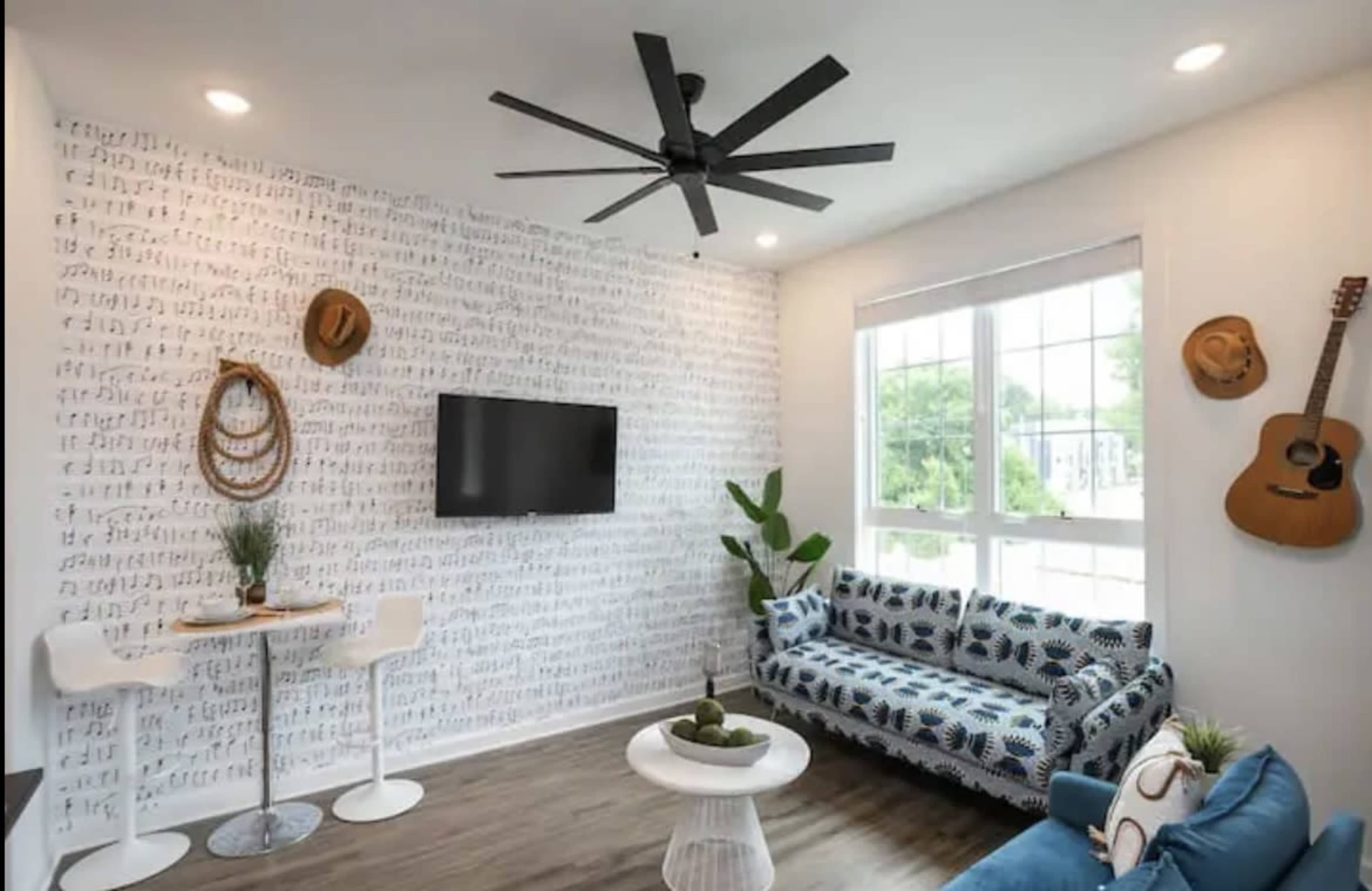 A modern living room with a patterned sofa, a television on the wall, a small dining table, and decorative items including hats and a guitar.