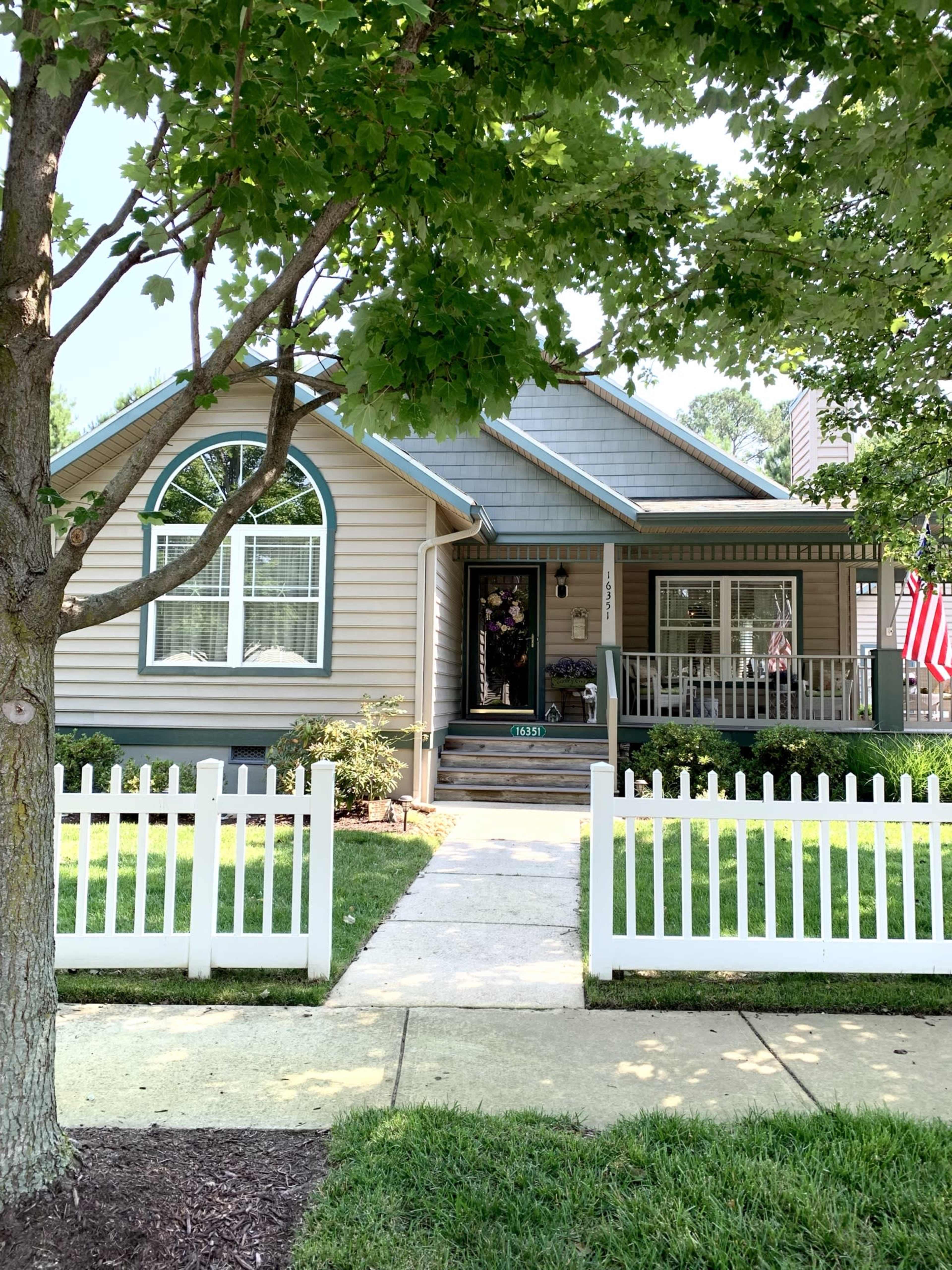 A single-story house with a front porch and white picket fence is framed by a tree and features an American flag hanging by the entrance.