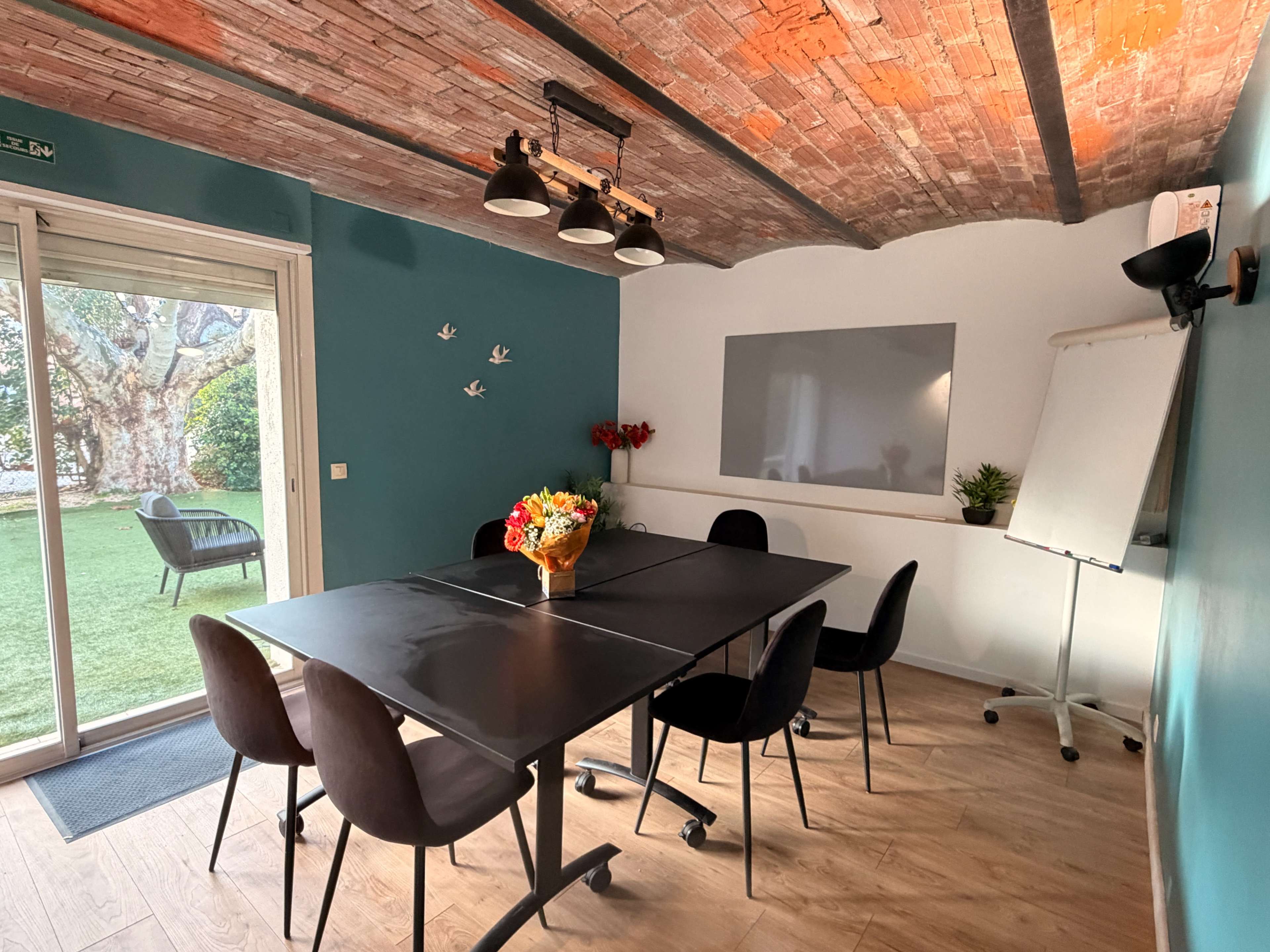 The image shows a meeting room with a large black table surrounded by black chairs, a whiteboard, and a window overlooking a garden.