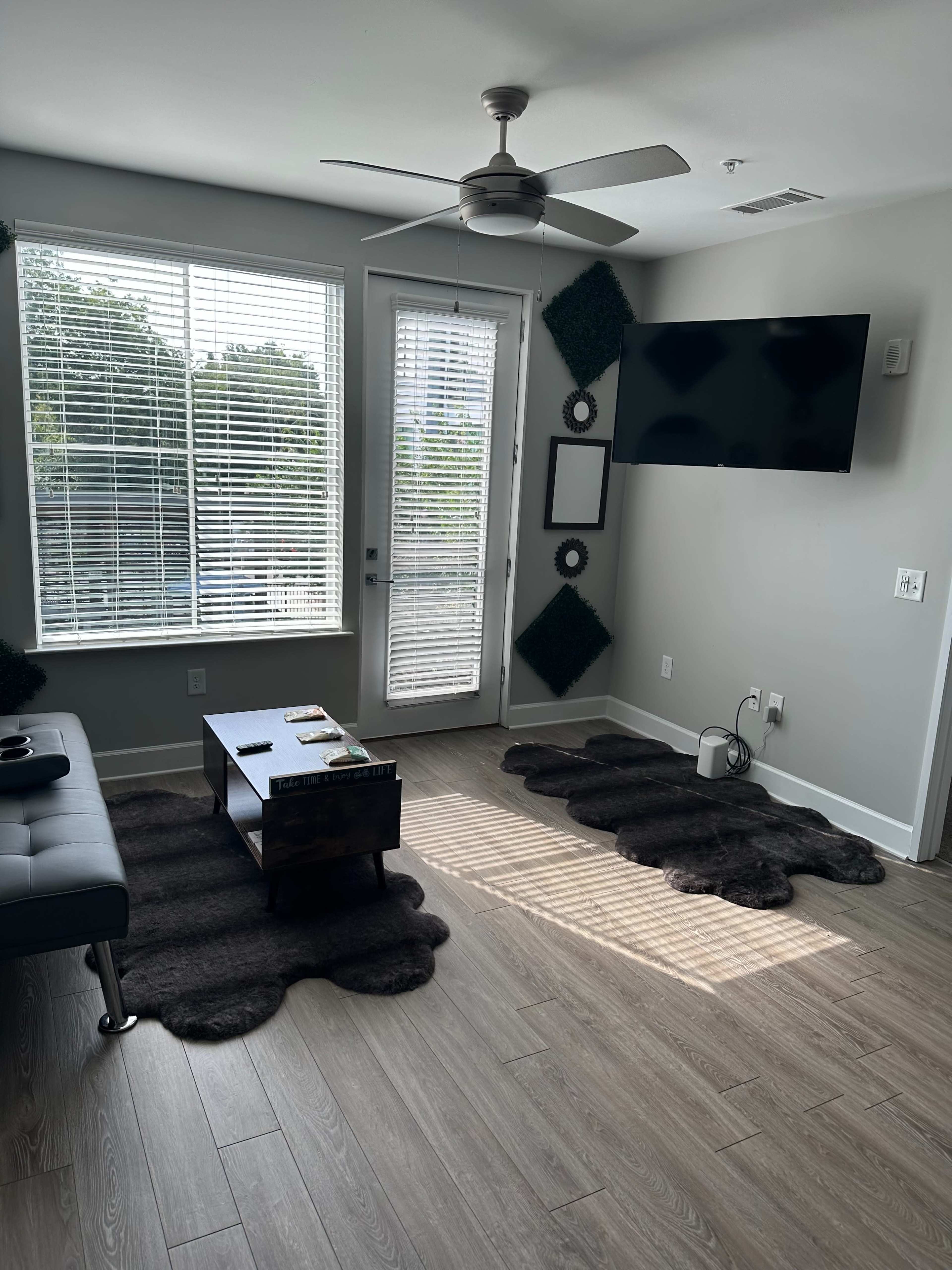 A modern living room features a dark rug, a coffee table, a wall-mounted TV, and large windows with blinds.