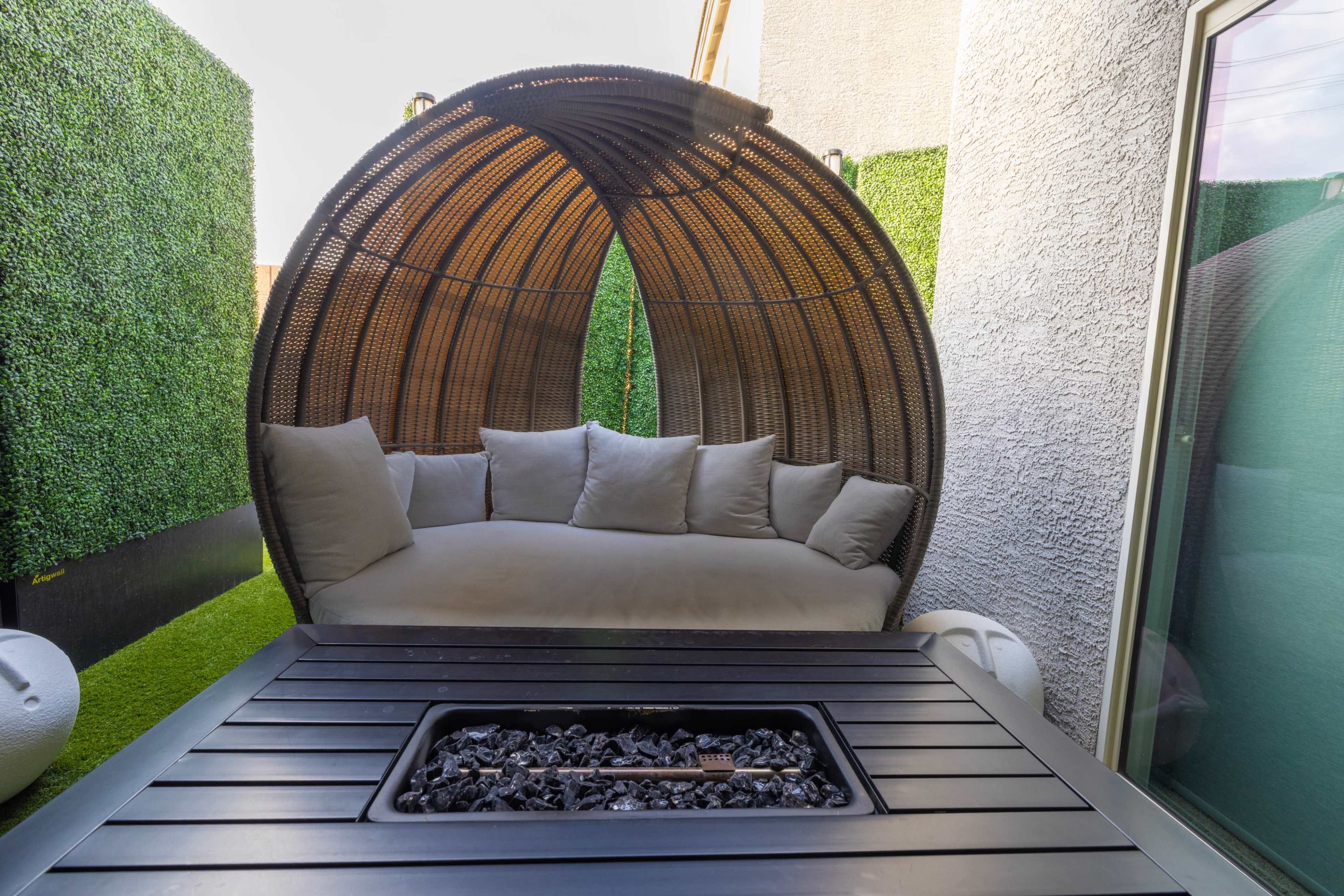 A circular woven lounge chair with cushions is positioned next to a rectangular fire table surrounded by artificial greenery.