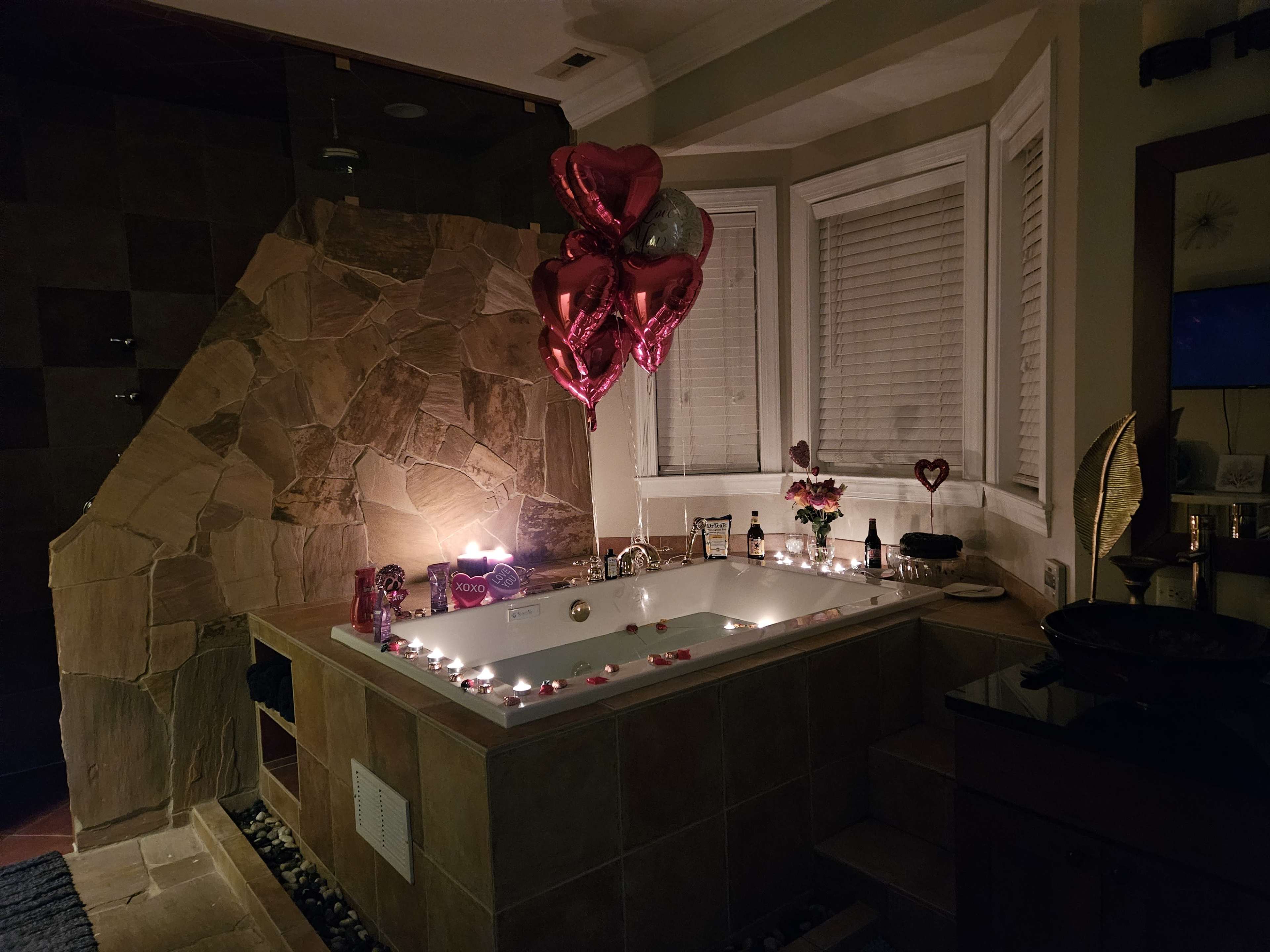 The image shows a dimly lit bathroom with a large bathtub decorated with rose petals and candles, accompanied by heart-shaped balloons.
