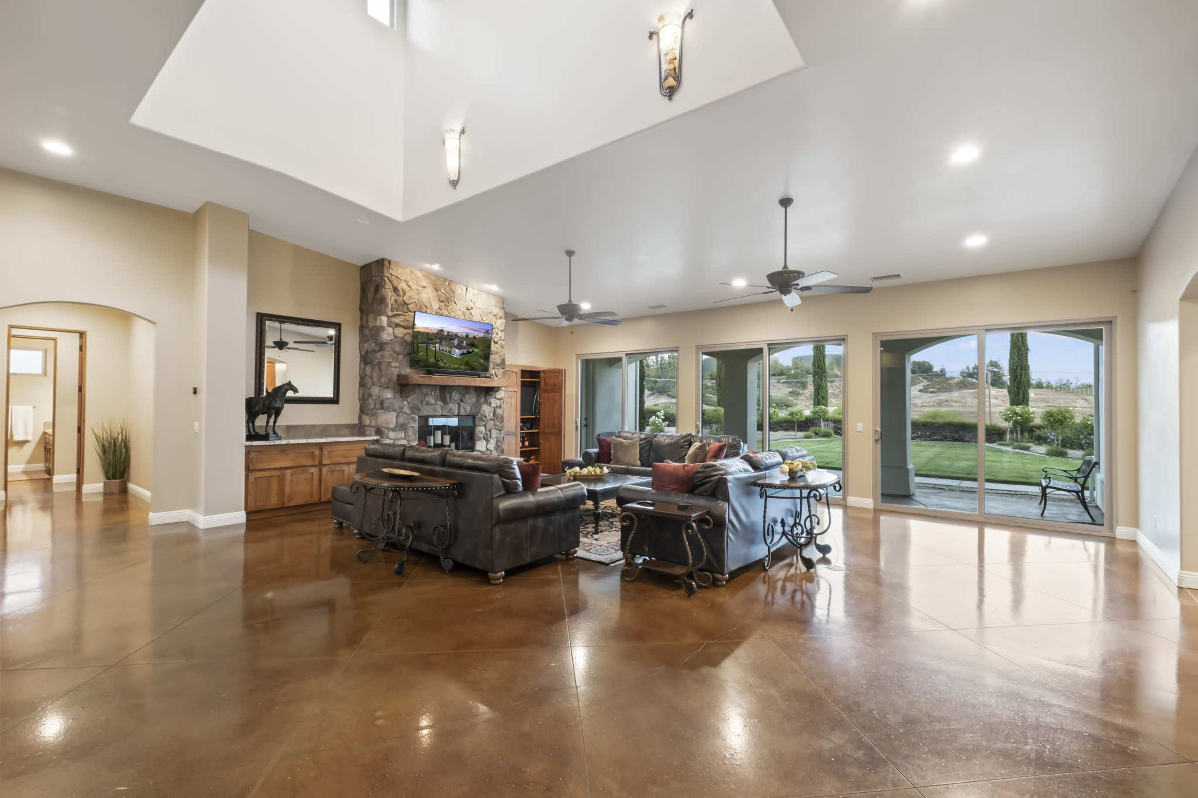 A spacious living room features a stone fireplace, large windows overlooking a backyard, and seating areas with leather sofas.