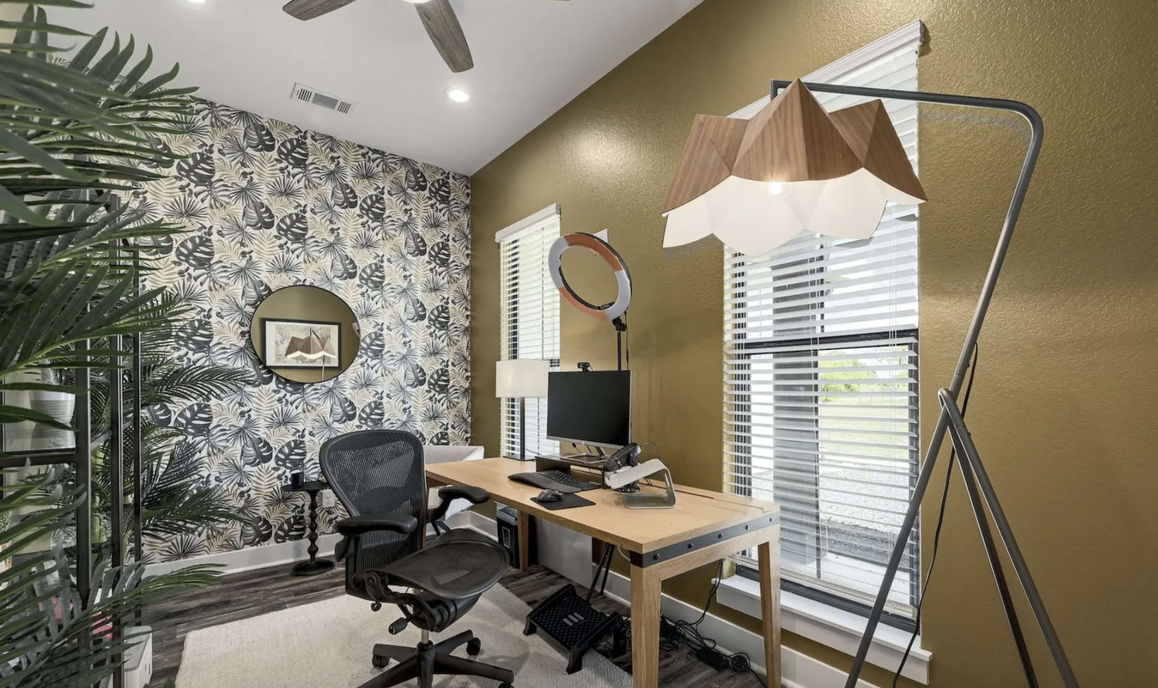 A home office features a wooden desk with a computer, two office chairs, a floor lamp, and a wall adorned with patterned wallpaper and a round mirror.