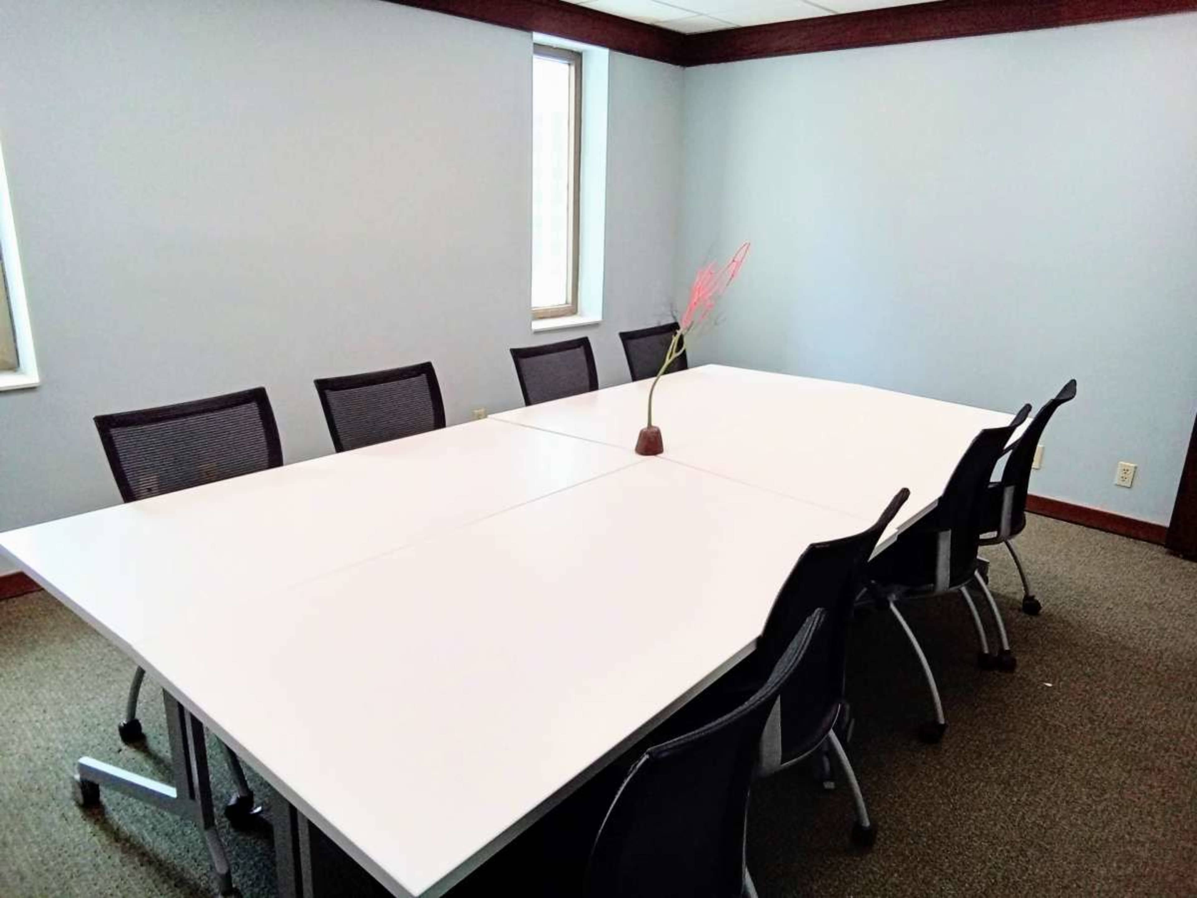 A conference room features a large white table surrounded by black chairs and a small vase with decorative branches in the center.