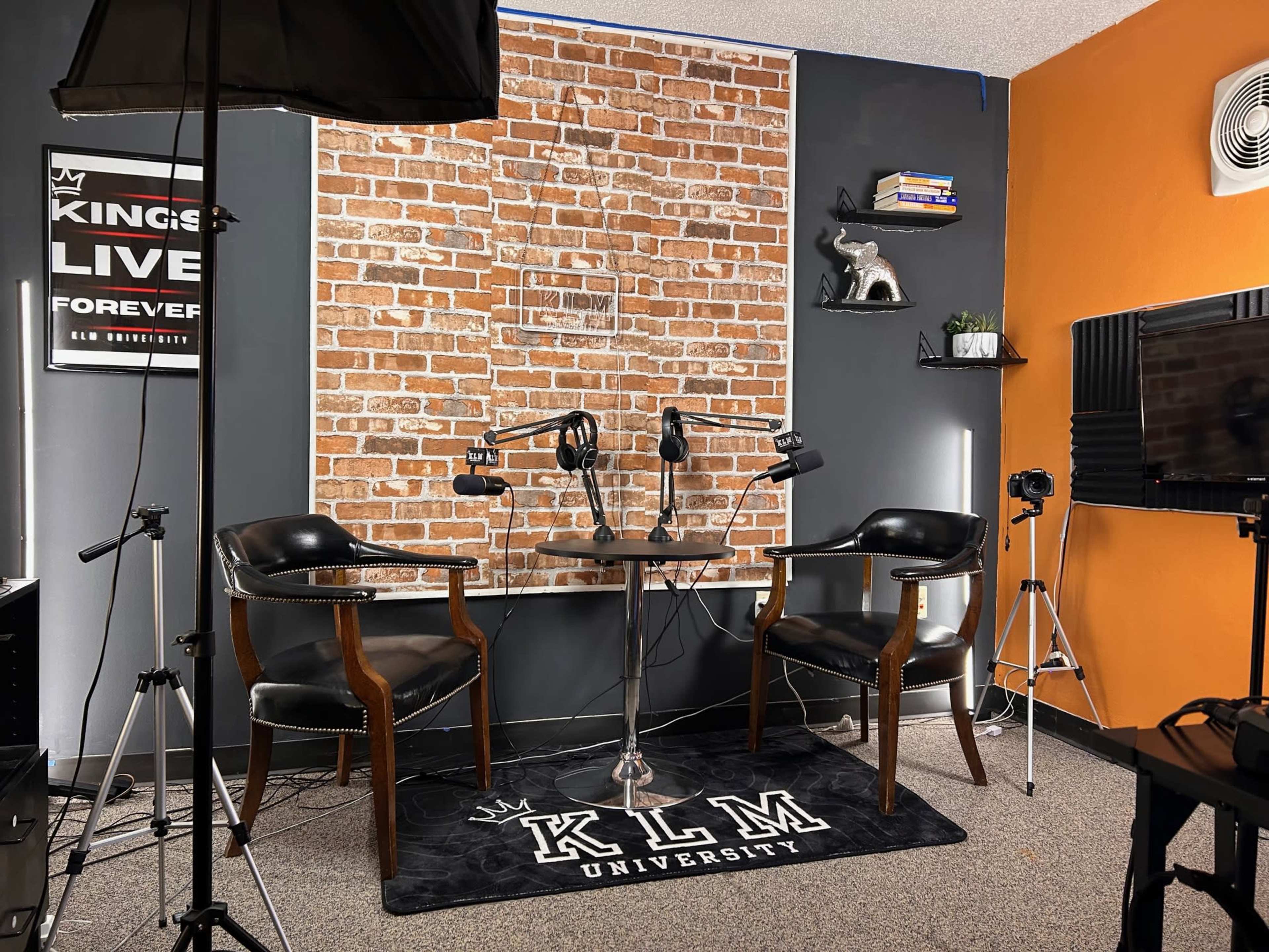 A podcast studio set with two chairs, a round table, and a brick wall backdrop, along with various lighting equipment and decorations.