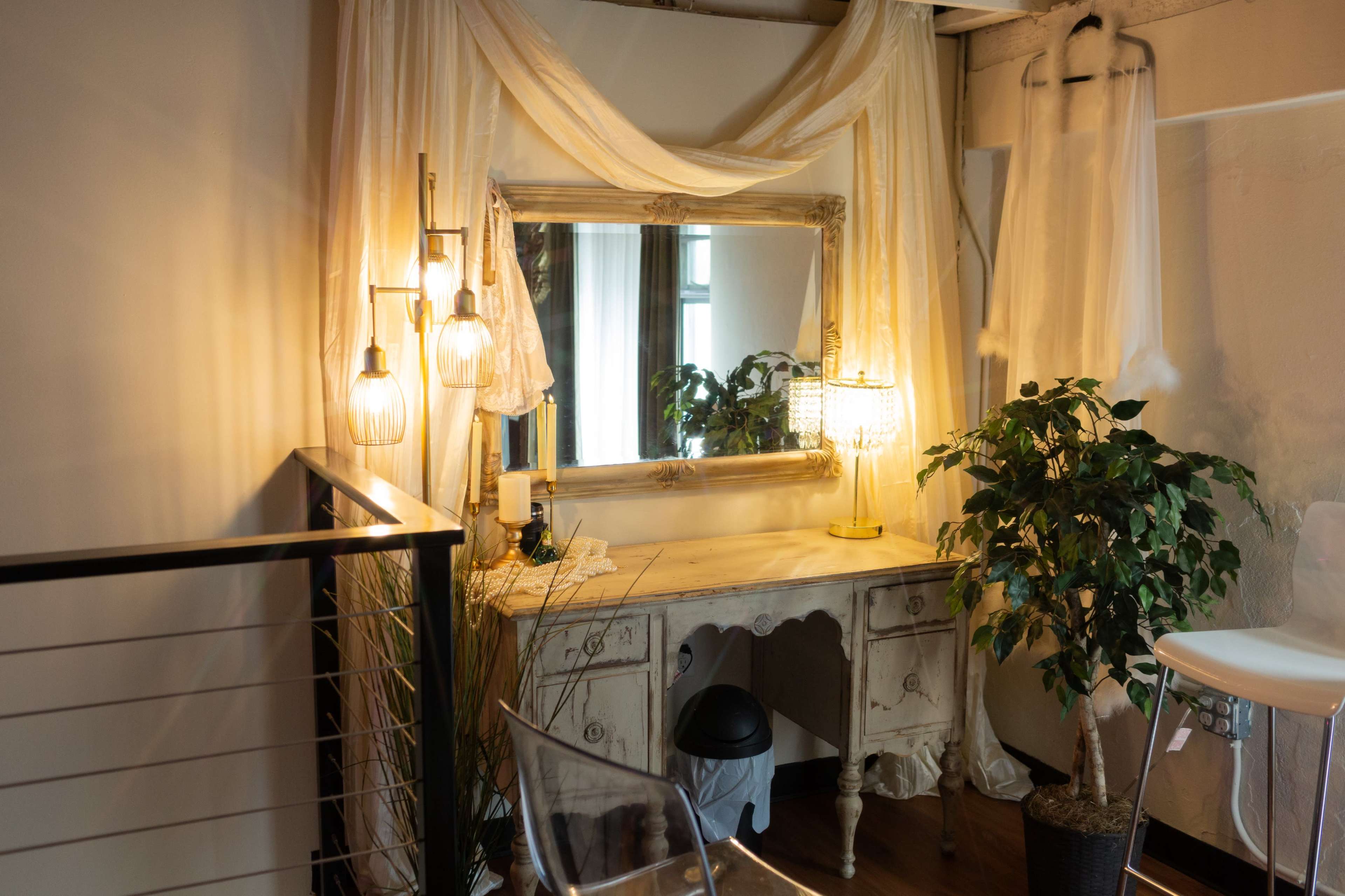 The image shows a vintage-style vanity table with a large mirror, surrounded by warm lighting fixtures and a potted plant in a softly decorated room.