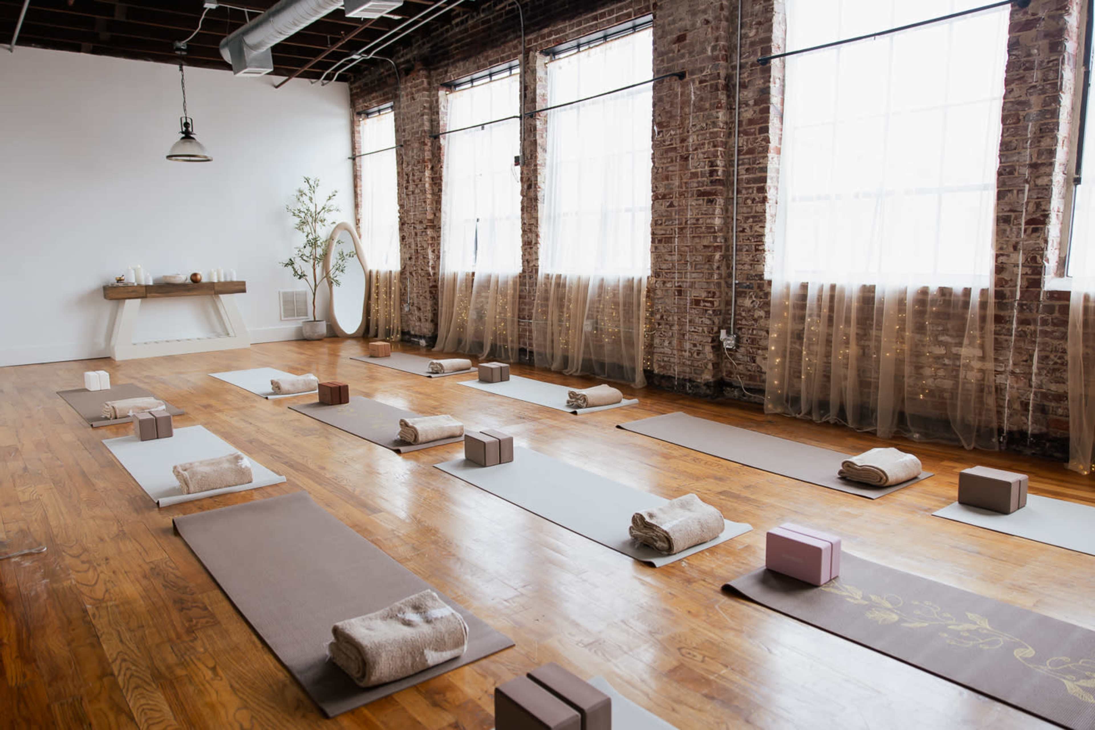 The image shows a spacious yoga studio with wooden floors, large windows, and yoga mats neatly arranged with props like blocks and towels.