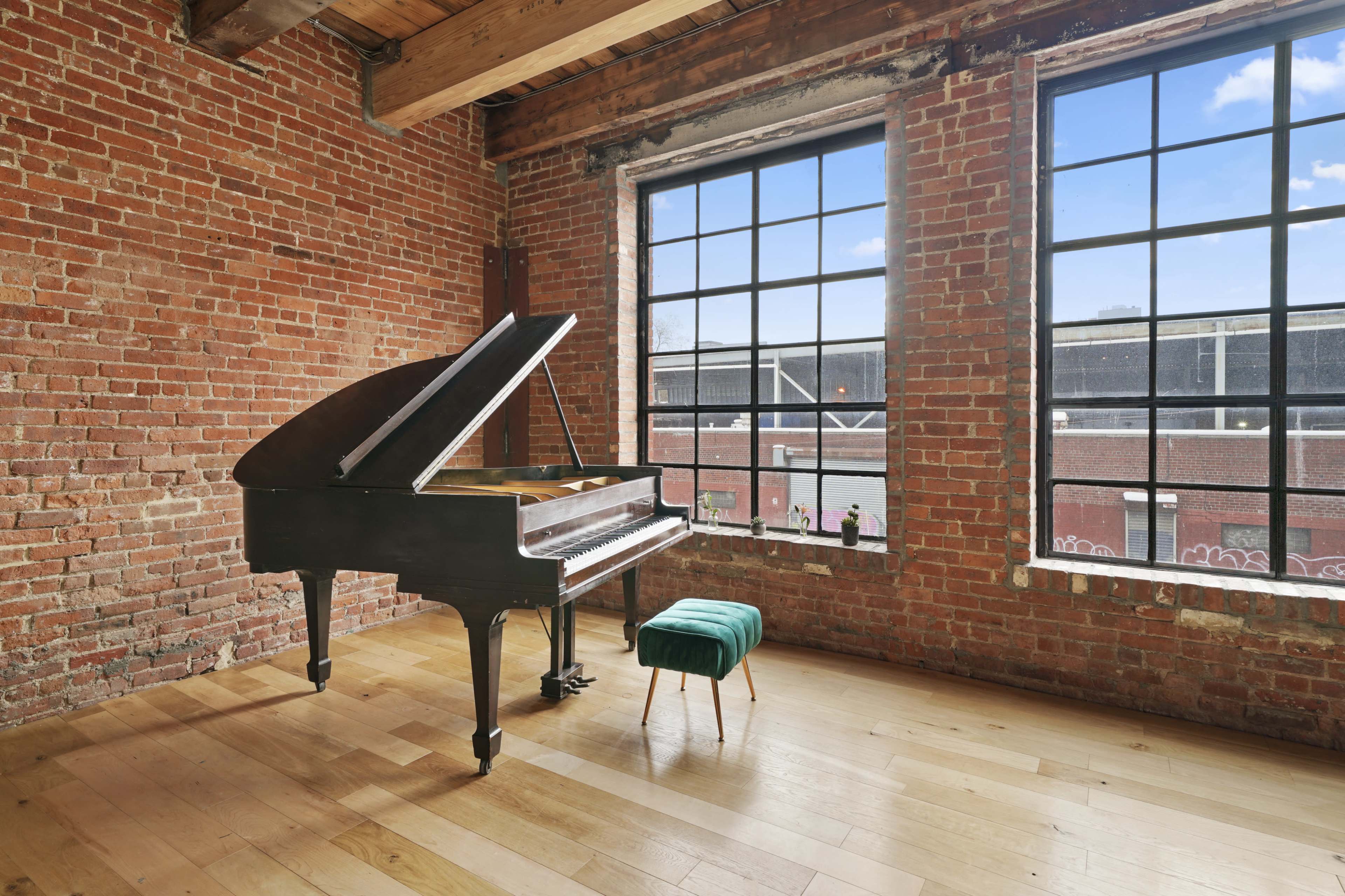 A grand piano sits next to large windows in a room characterized by brick walls and wooden flooring.
