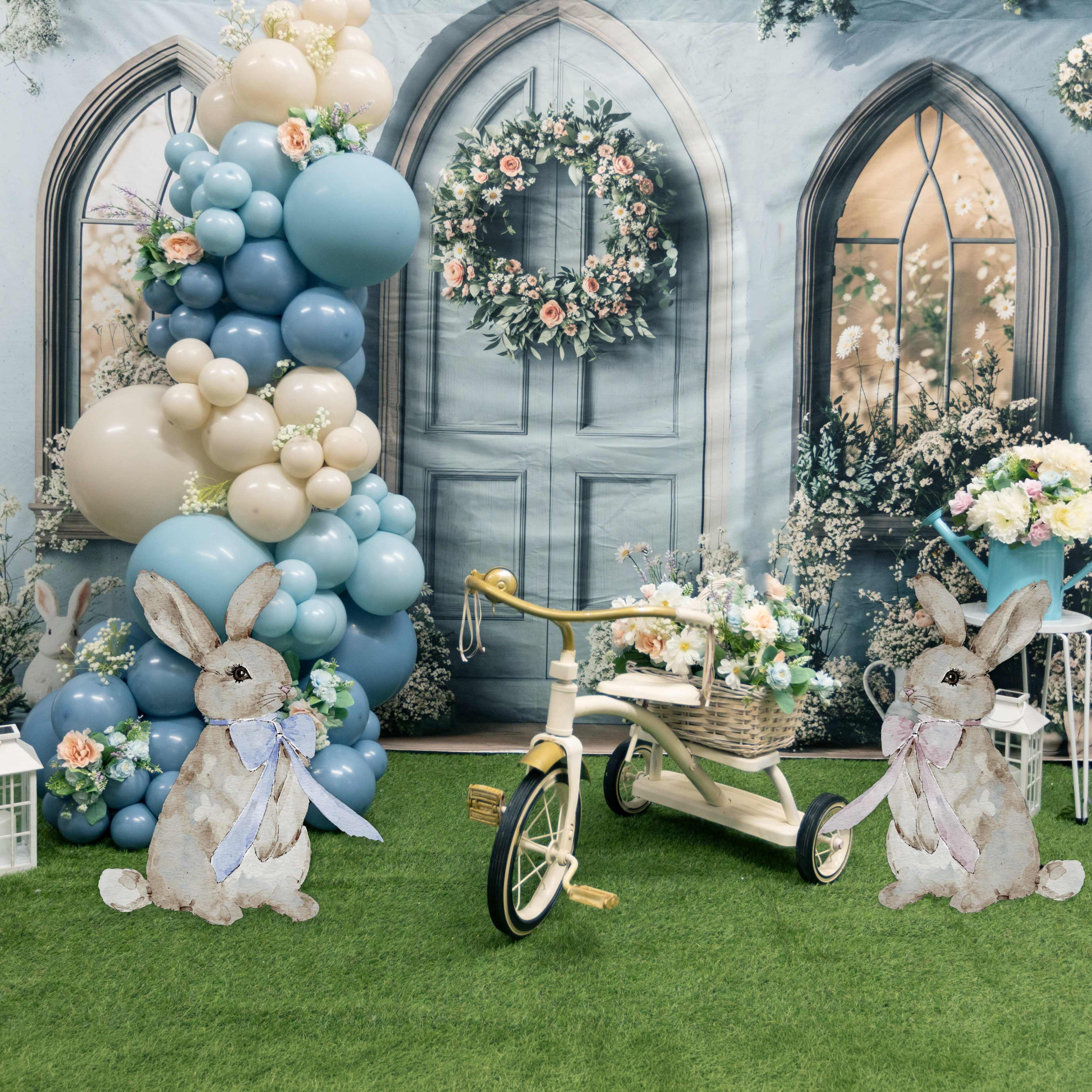 The scene features a decorative setup with two cardboard bunny figures, a vintage-style tricycle, and a backdrop of balloons and floral arrangements.