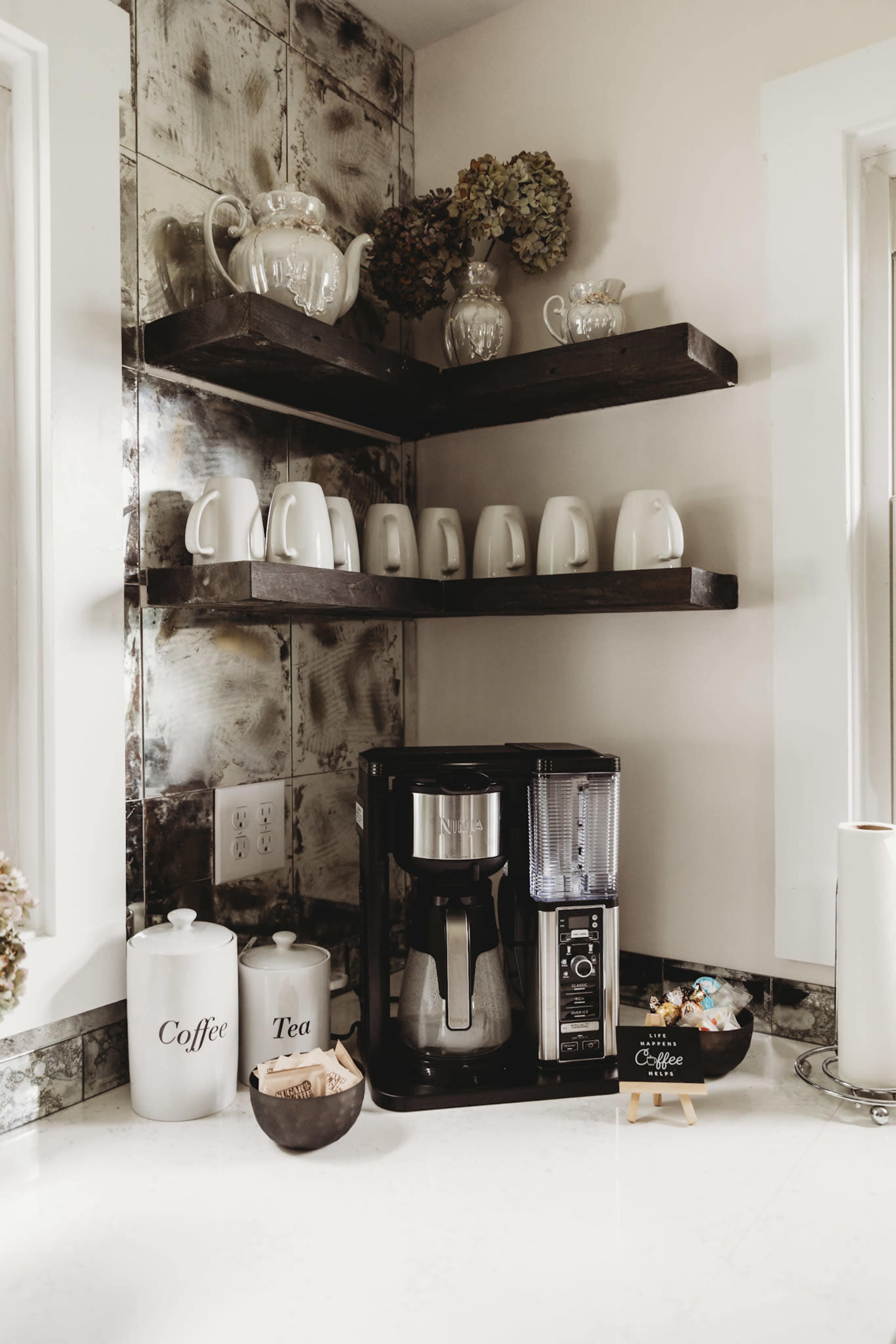 A coffee maker sits on a countertop beneath open shelves holding white mugs and decorative teapots.