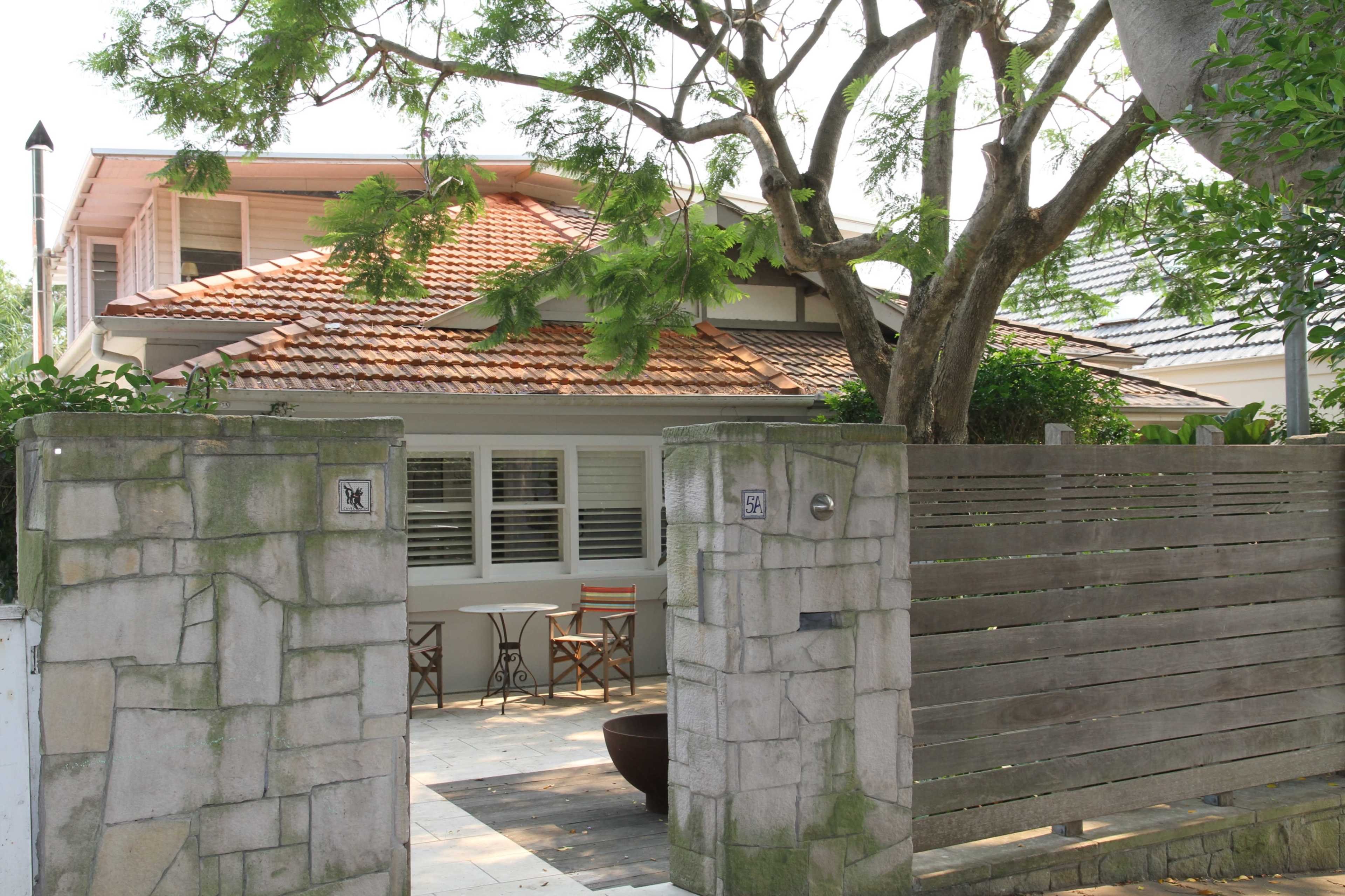 A single-story house with a tiled roof is visible through a stone and wooden gate, accompanied by outdoor seating beneath a tree.