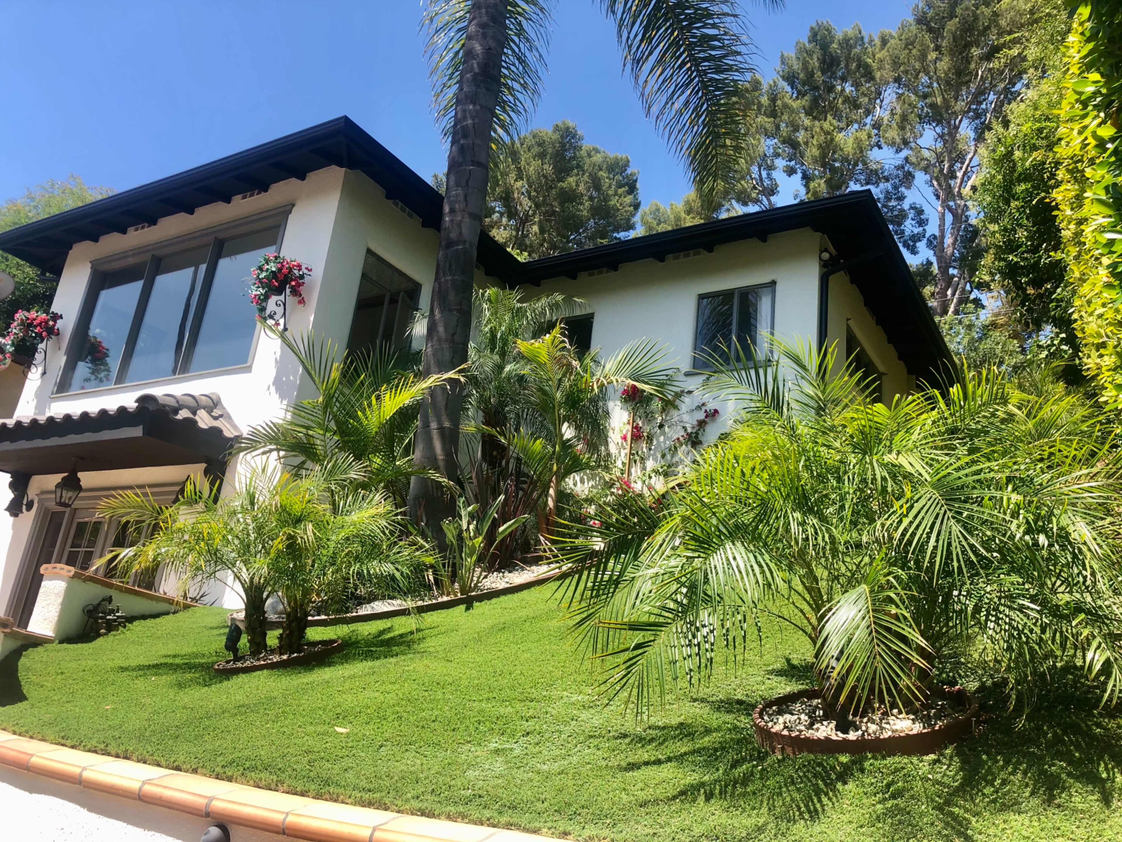 The image shows a two-story white house with a sloped green lawn, adorned with palm trees and flowering plants on the exterior.