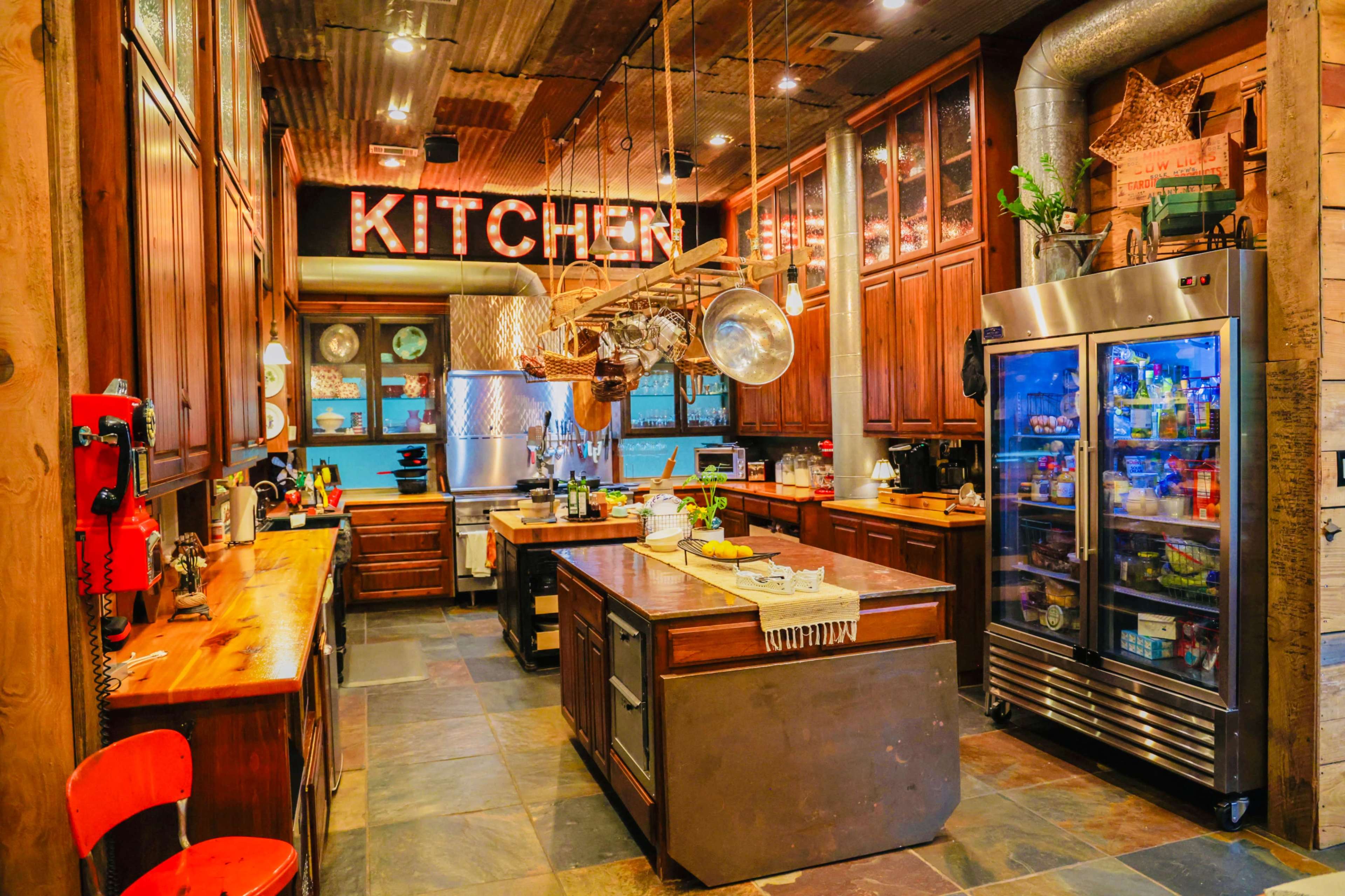 A rustic kitchen features wooden cabinets, a central island, and a large refrigerator beside a glass-fronted pantry.