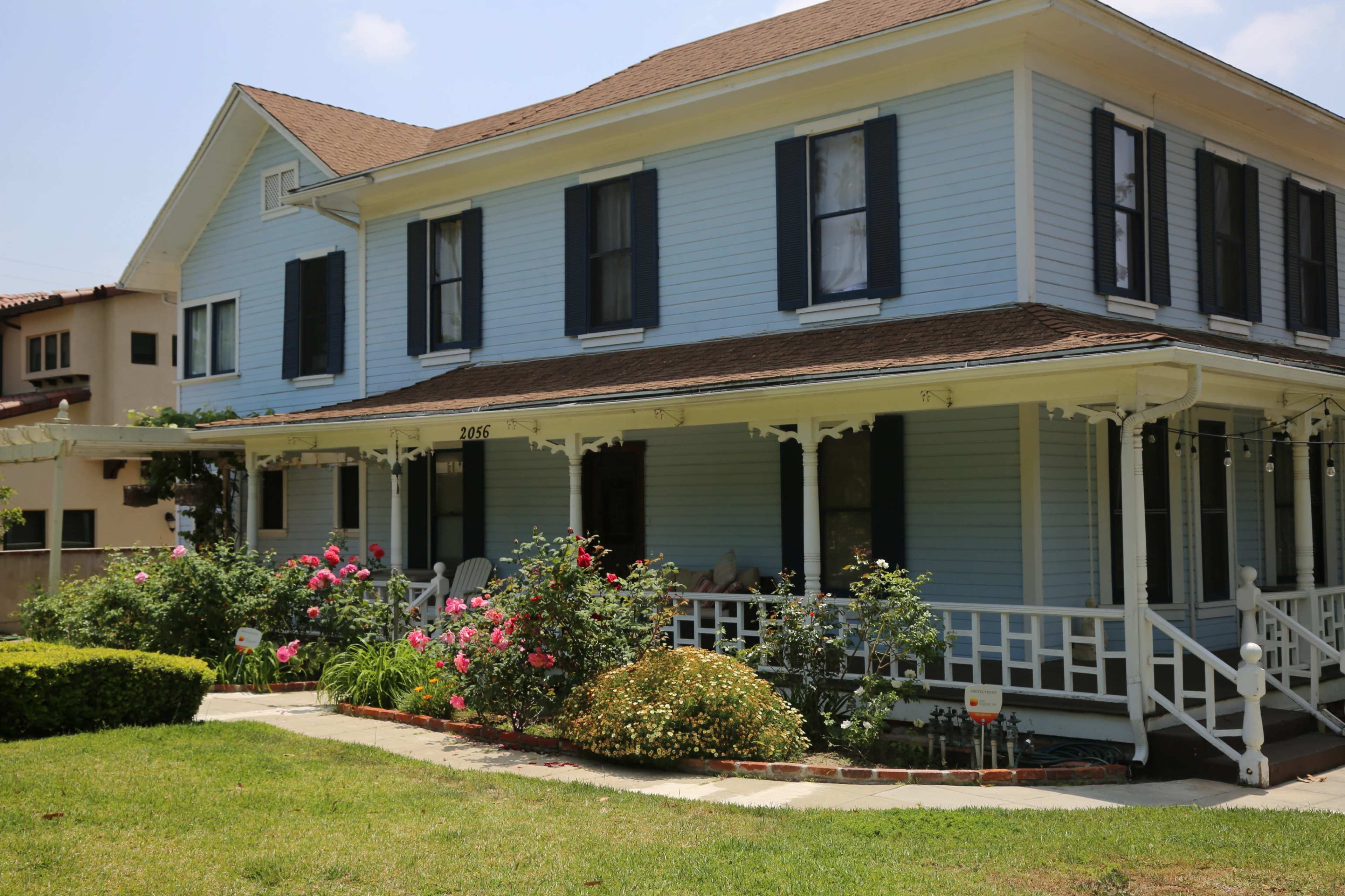 A light blue two-story house with black shutters and a front porch, surrounded by a well-maintained garden with pink flowers.