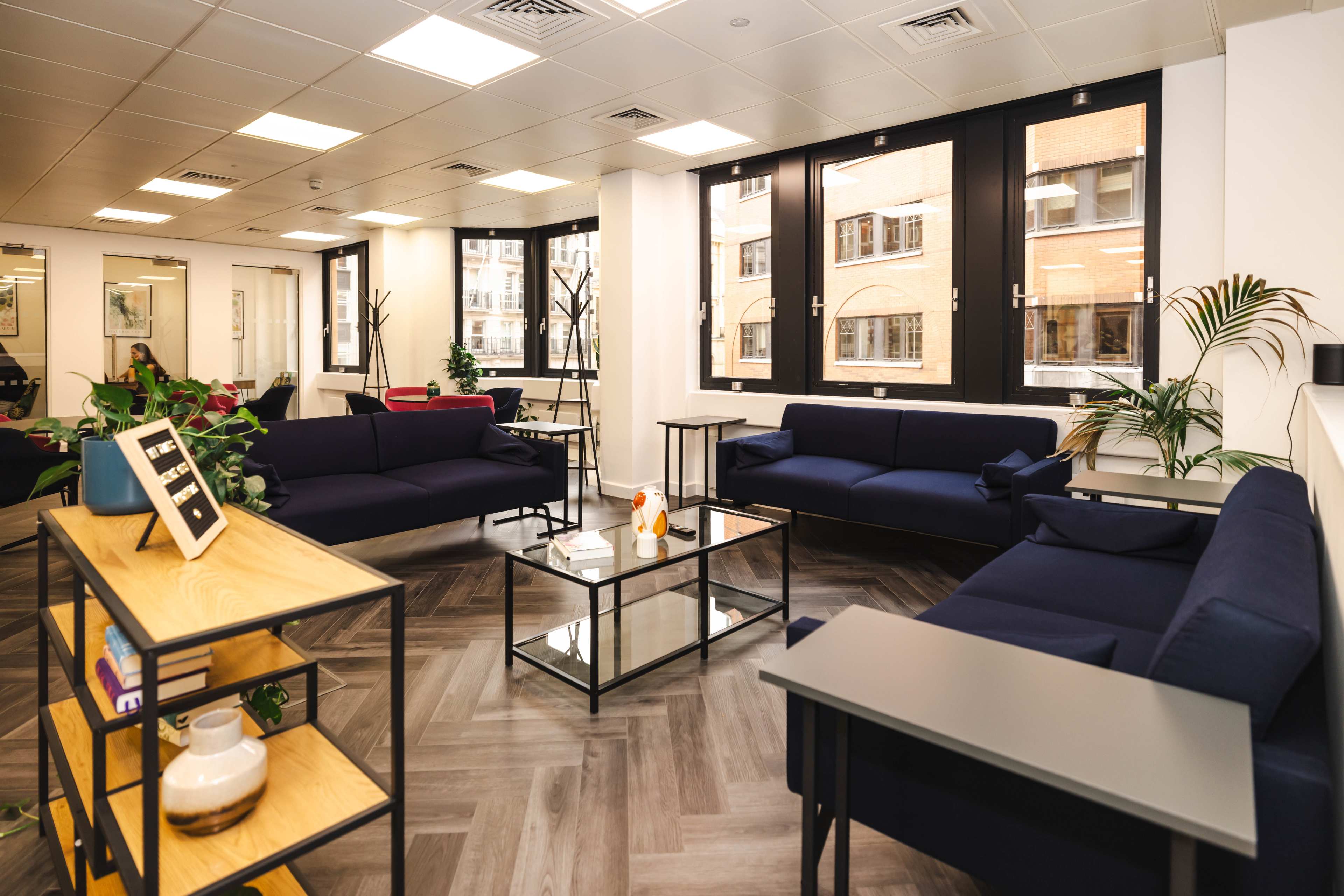 The image shows a modern office lounge area with blue sofas, glass coffee tables, and plants, illuminated by natural light from large windows.