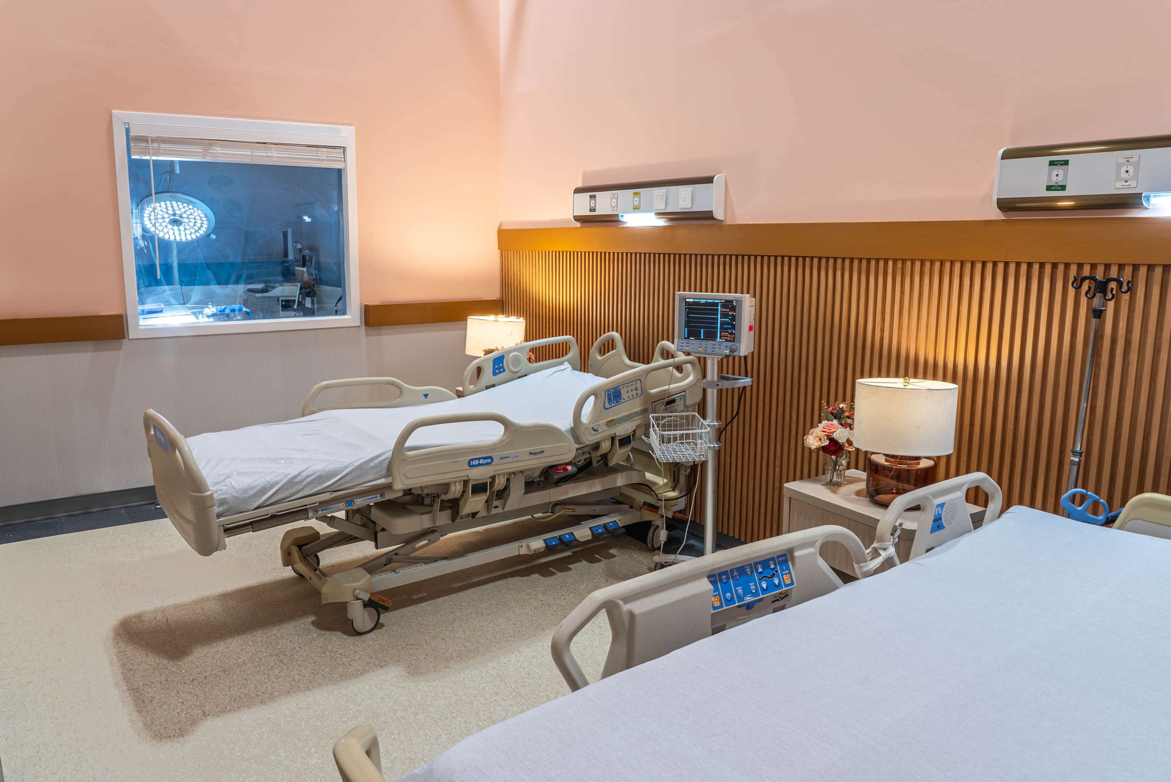The image shows a hospital room with a medical bed, monitoring equipment, and two additional beds in a clean, well-lit environment.