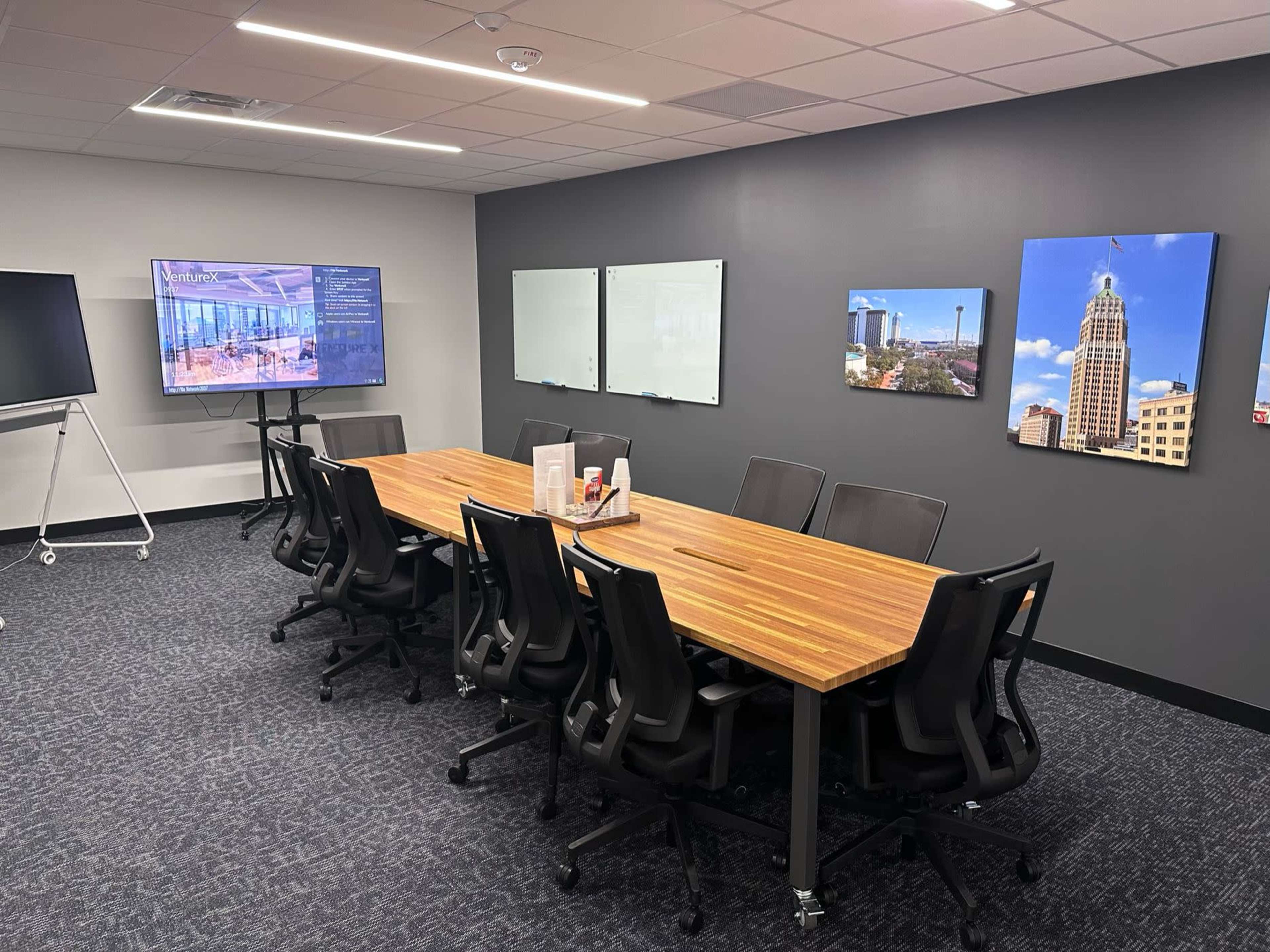 A modern conference room features a large wooden table surrounded by office chairs, with screens and whiteboards mounted on the walls.