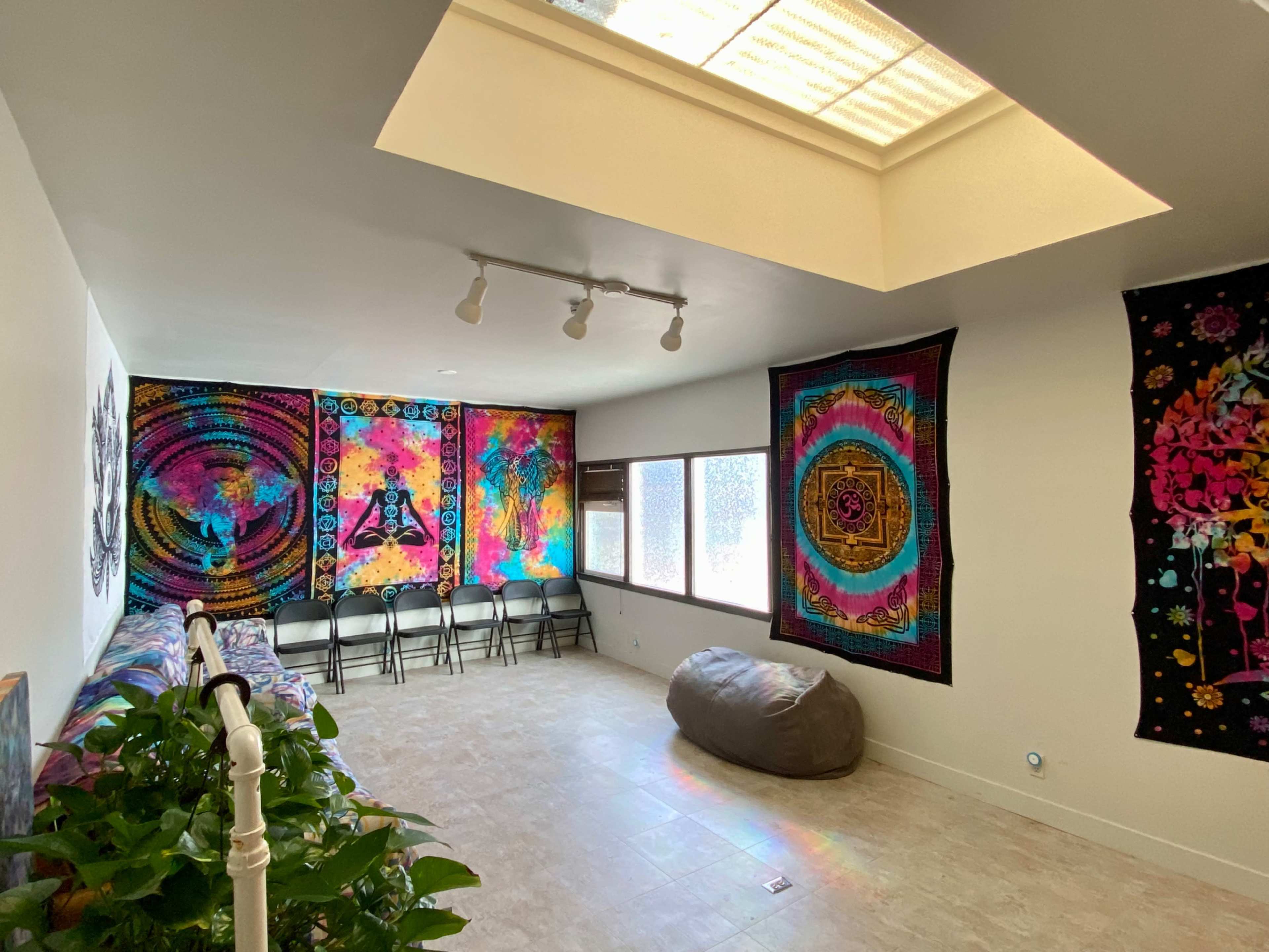 The room features colorful tapestry wall art, a row of folding chairs, and a large bean bag on a tiled floor illuminated by natural light from a skylight.
