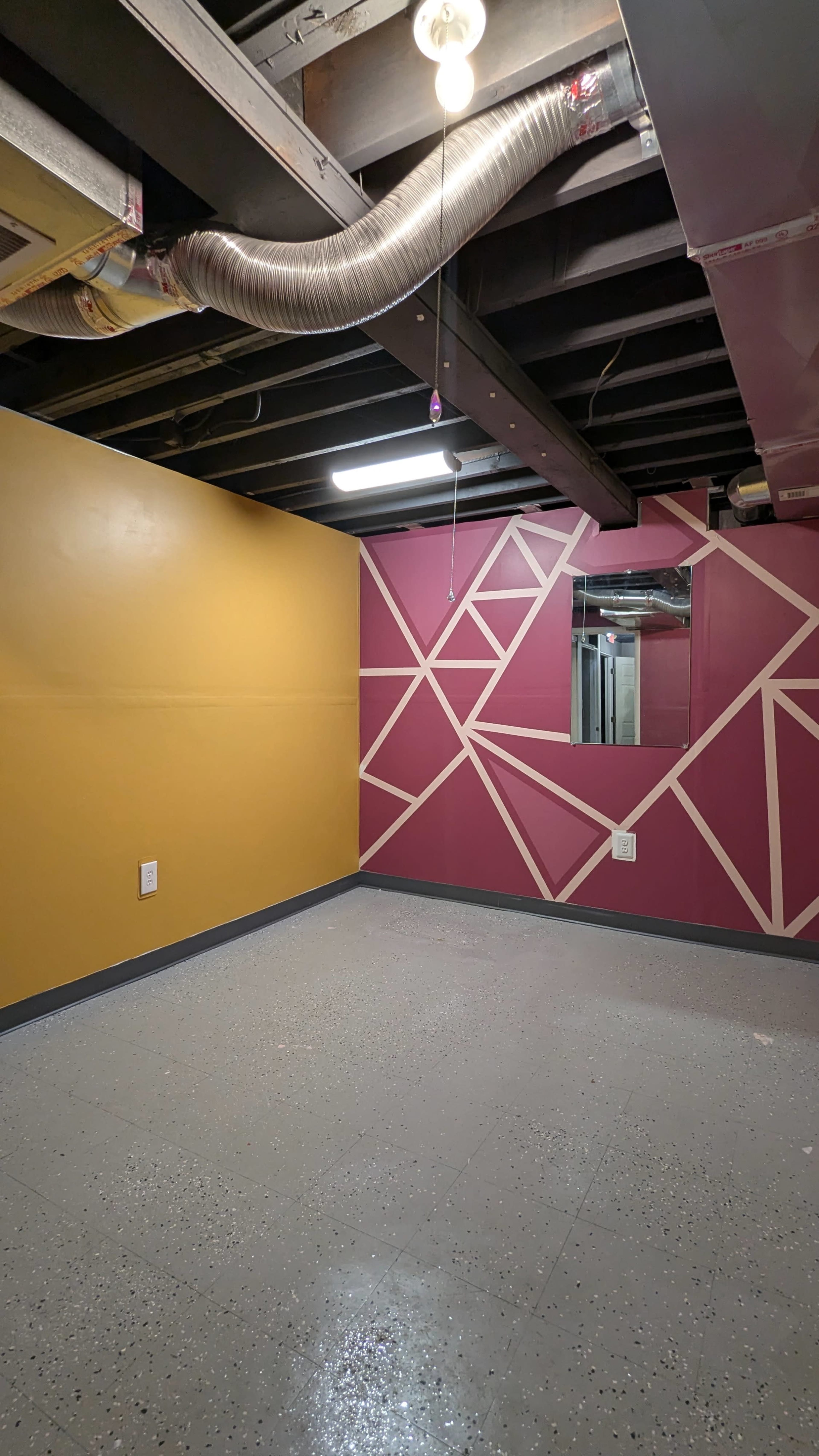The image shows a basement room with one wall painted yellow, another wall featuring a pink geometric design, and a mirror mounted on the pink wall.