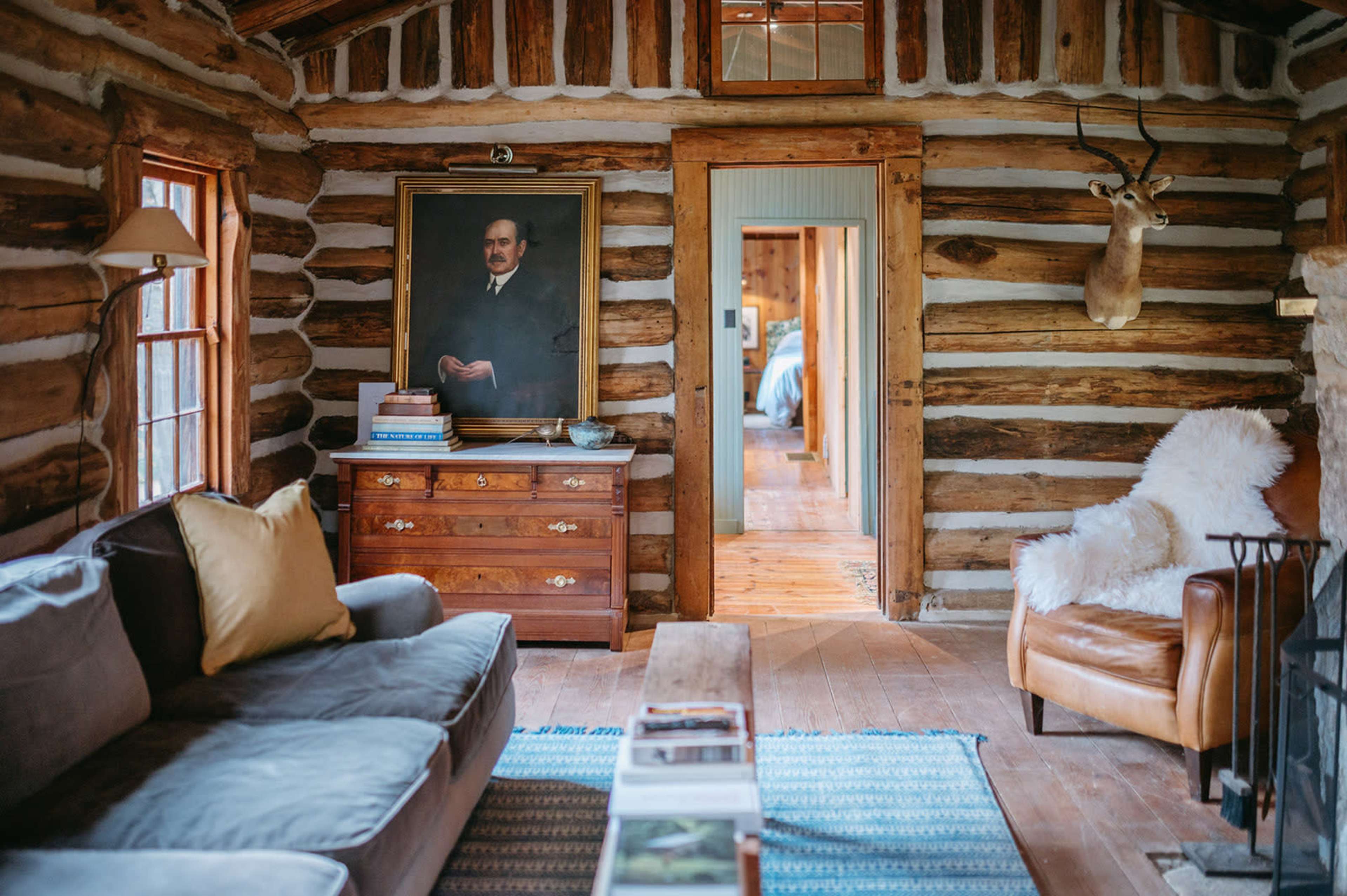 The image shows a cozy log cabin interior featuring a sofa, a wooden chest of drawers with a portrait above it, and a mounted deer head on the wall.