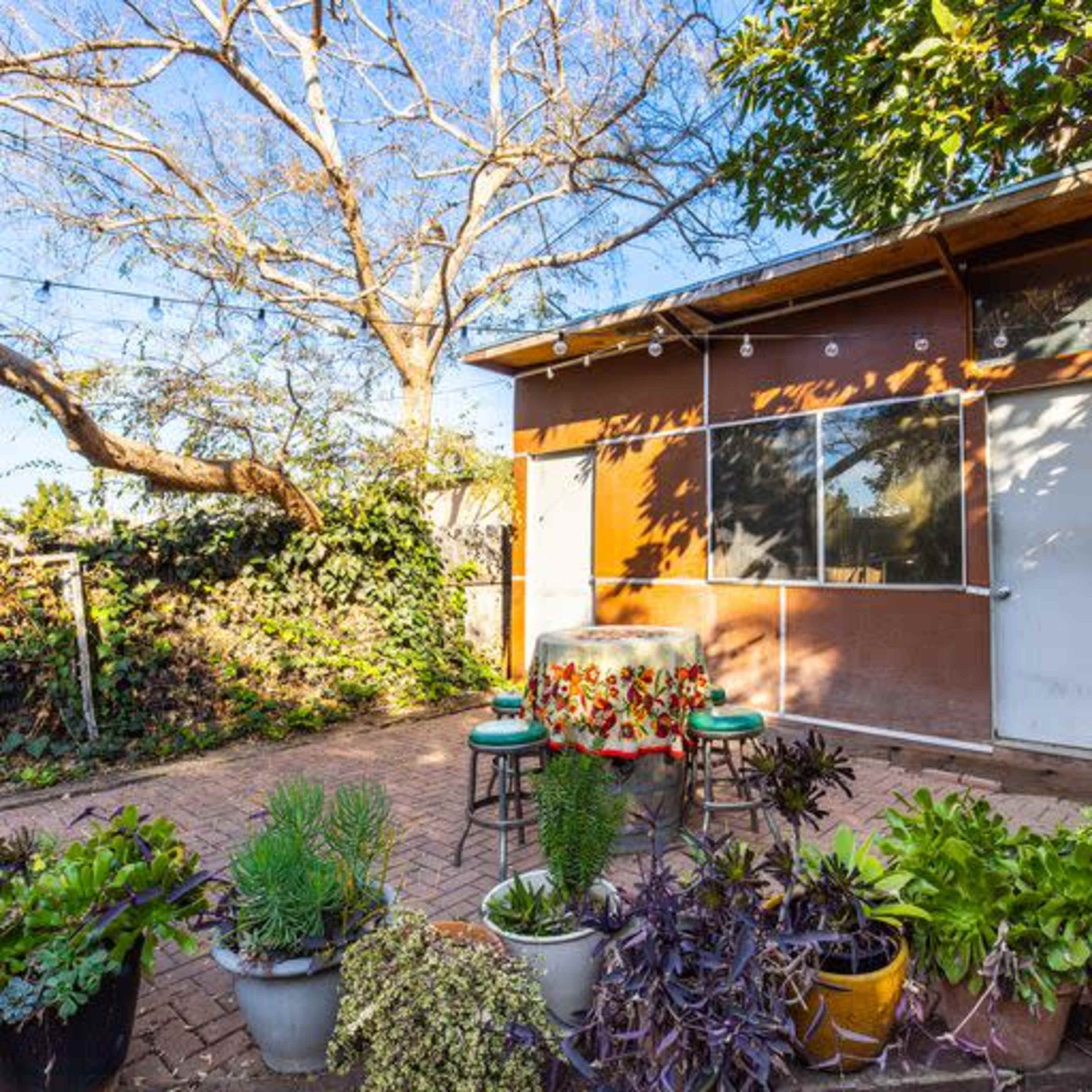 A sunlit outdoor patio area with a wooden structure, surrounded by plants and a table set for dining.