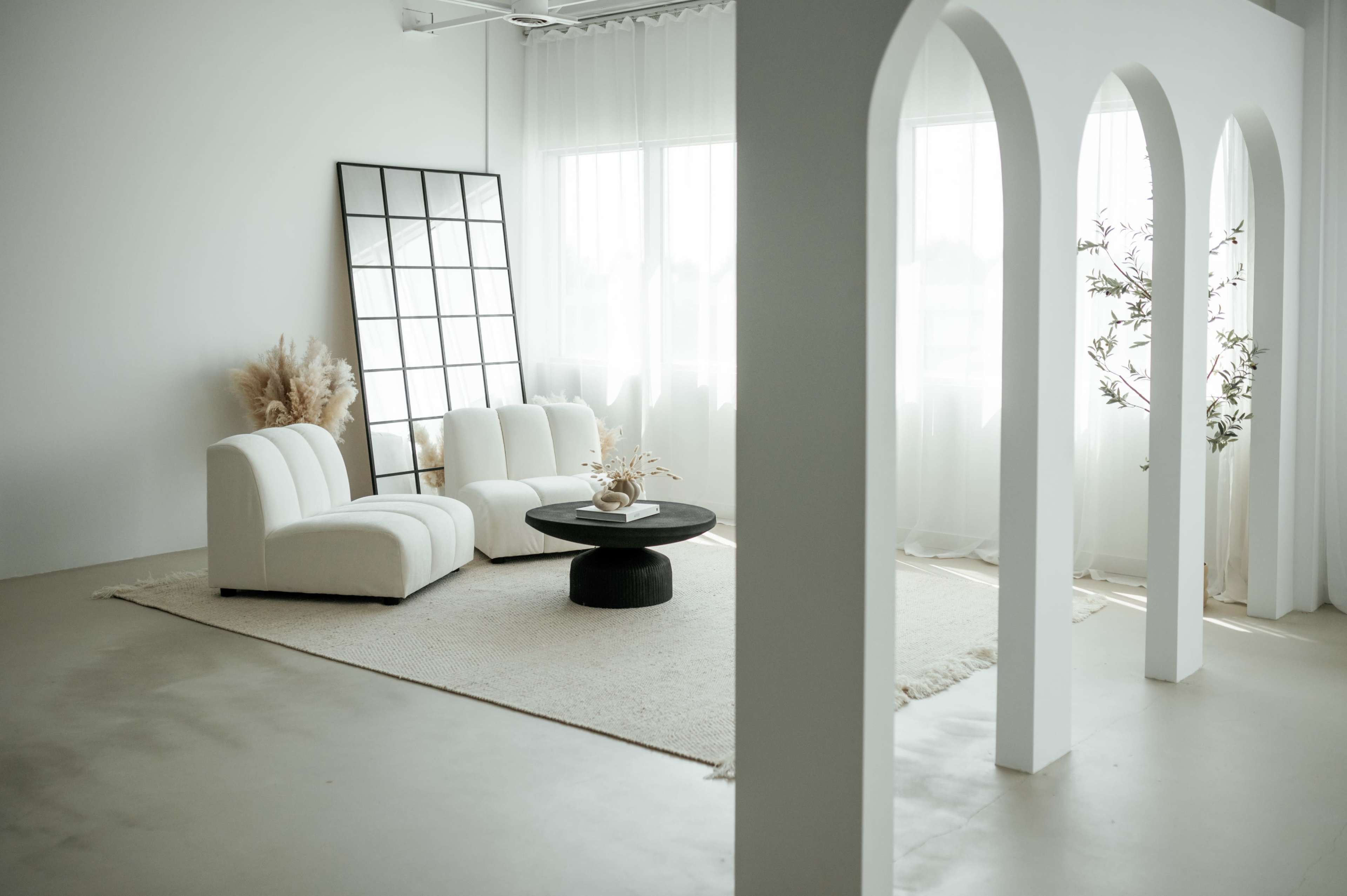 A minimalist living room features white furniture, a large mirror, and a round black coffee table, surrounded by sheer curtains and a light-colored rug.