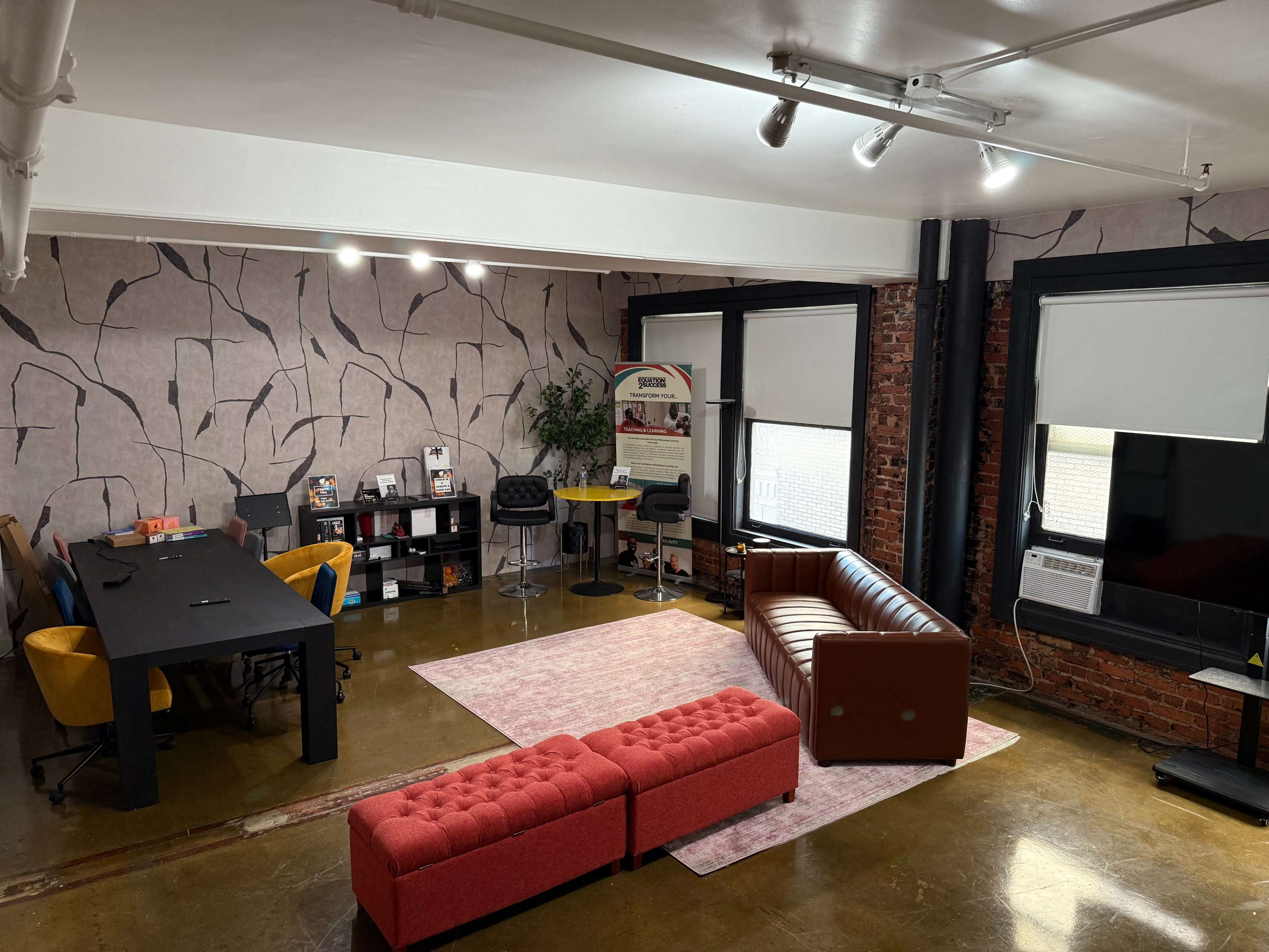 The image shows a modern office space featuring a large black table, colorful chairs, a brown leather sofa, a pink rug, and a wall with a textured design.