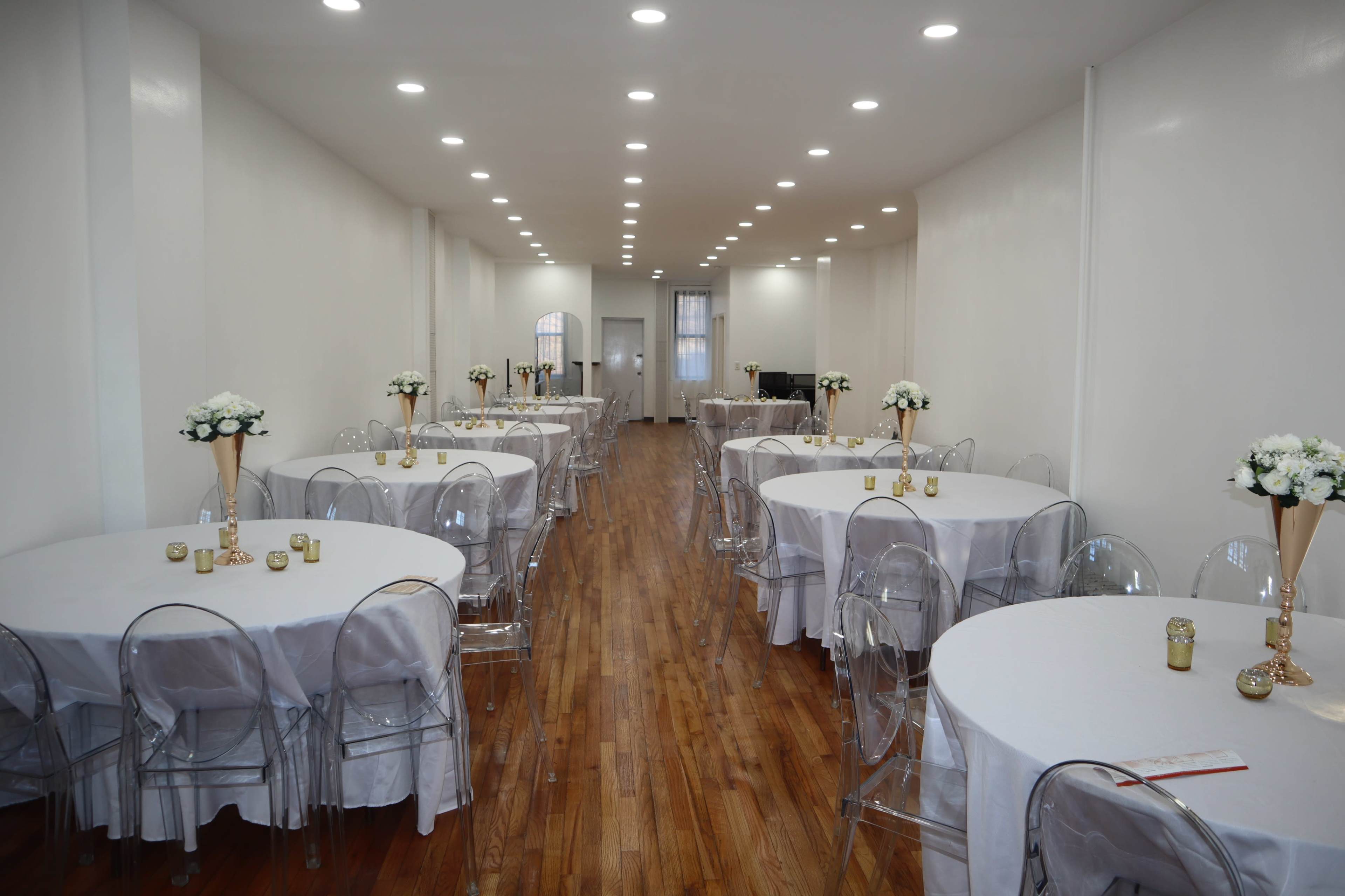 A brightly lit indoor space arranged with several round tables covered in white tablecloths, each adorned with flower centerpieces and gold candle holders, along a long wooden floor.