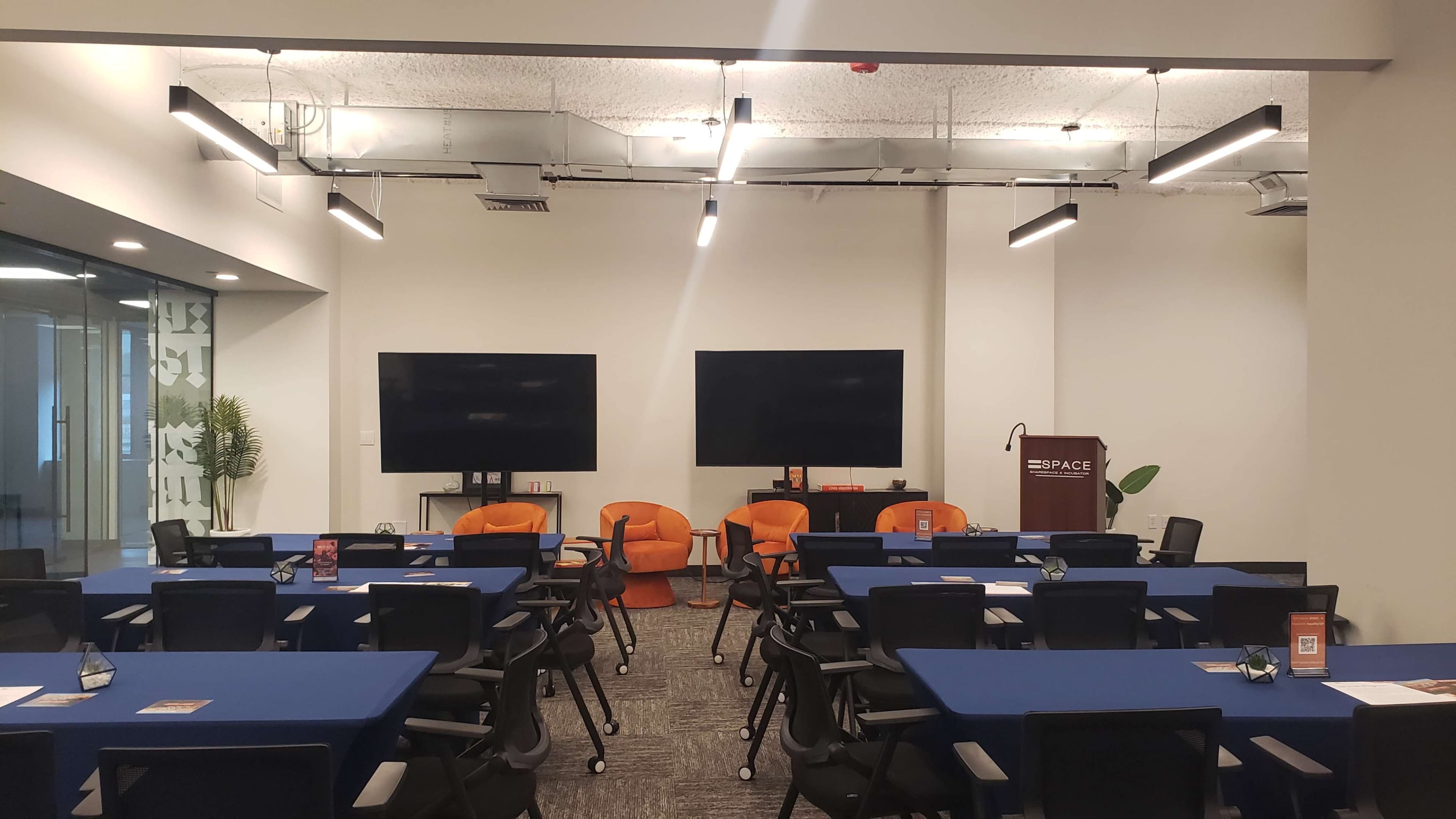 The image shows a modern conference room with blue tablecloths on several tables, two large screens on the front wall, and orange chairs arranged in front of the screens.