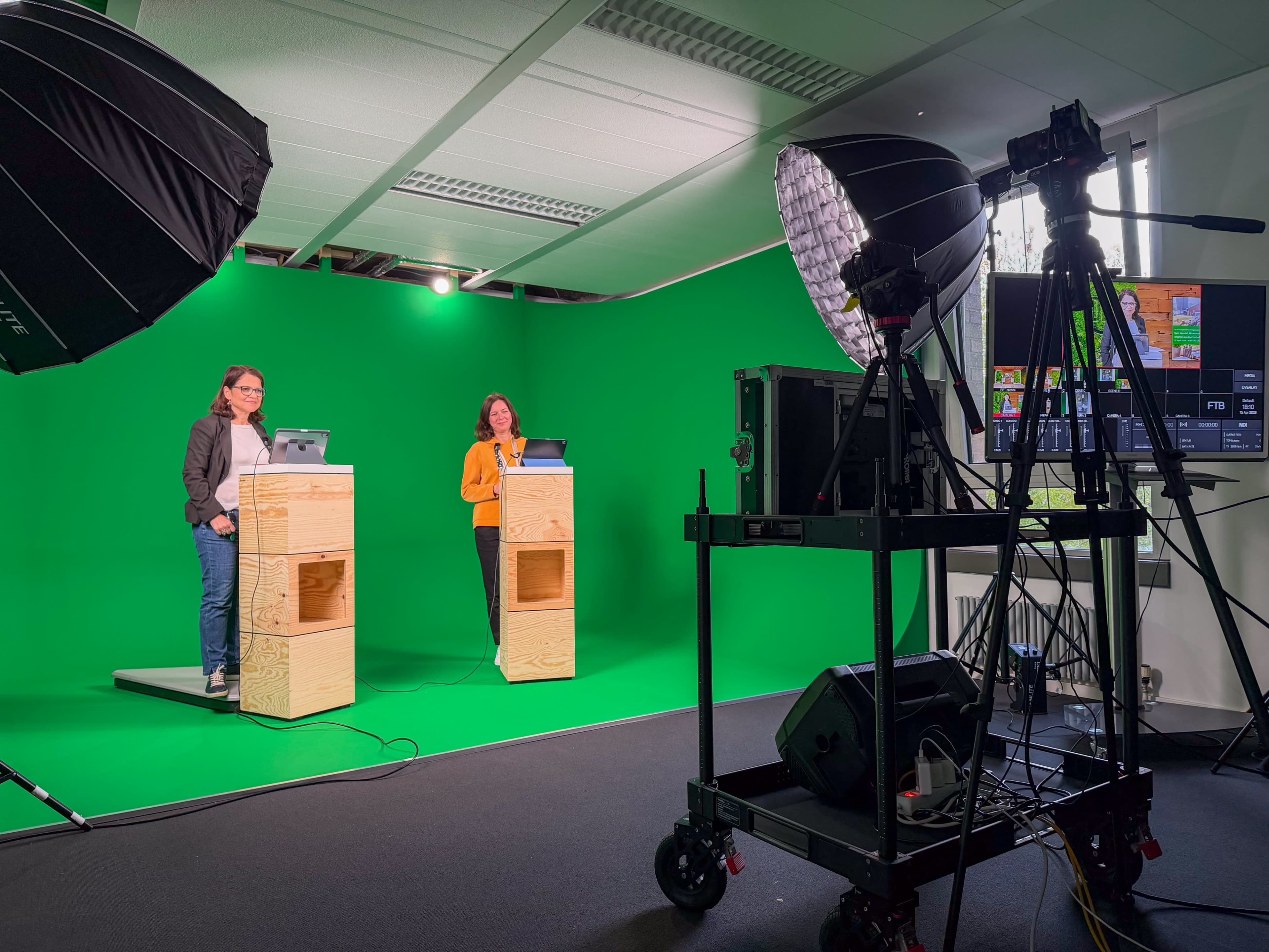 Two presenters stand behind wooden podiums in front of a green screen setup, equipped with cameras and lighting equipment for a studio production.