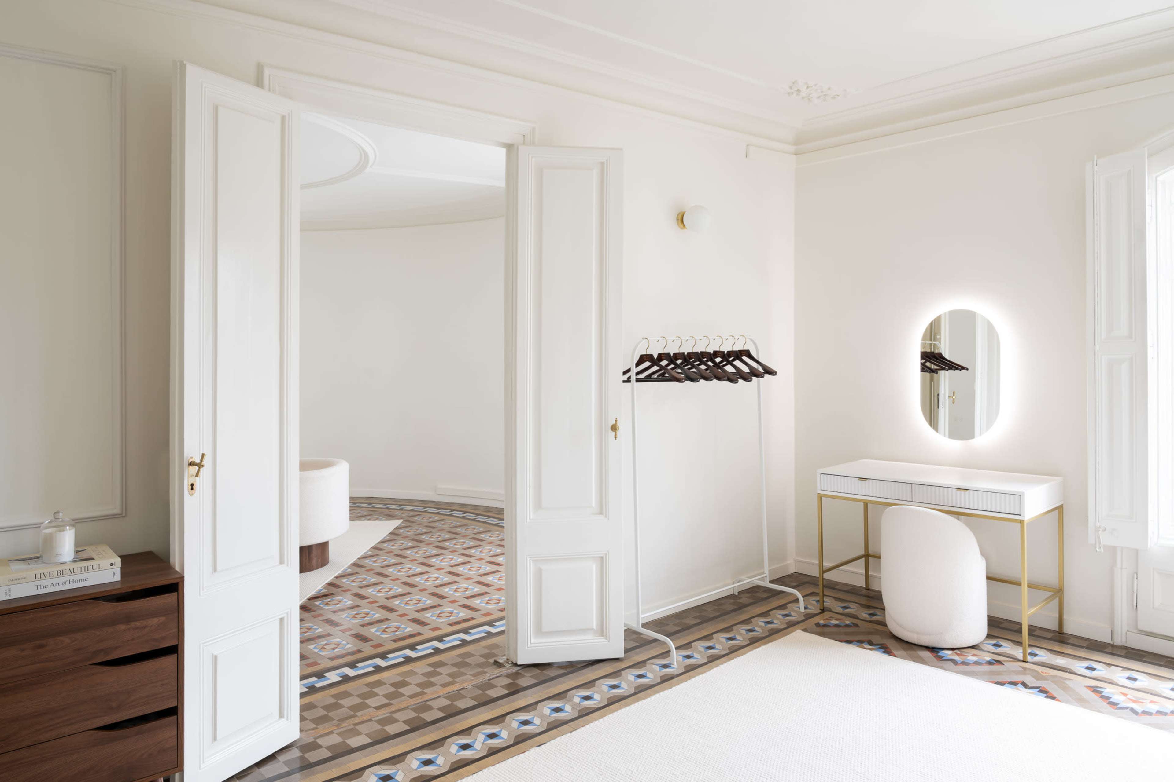 The image shows a minimalist interior room with light-colored walls, a patterned floor, a dressing table with a mirror, a clothing rack, and large windows that allow natural light to enter.