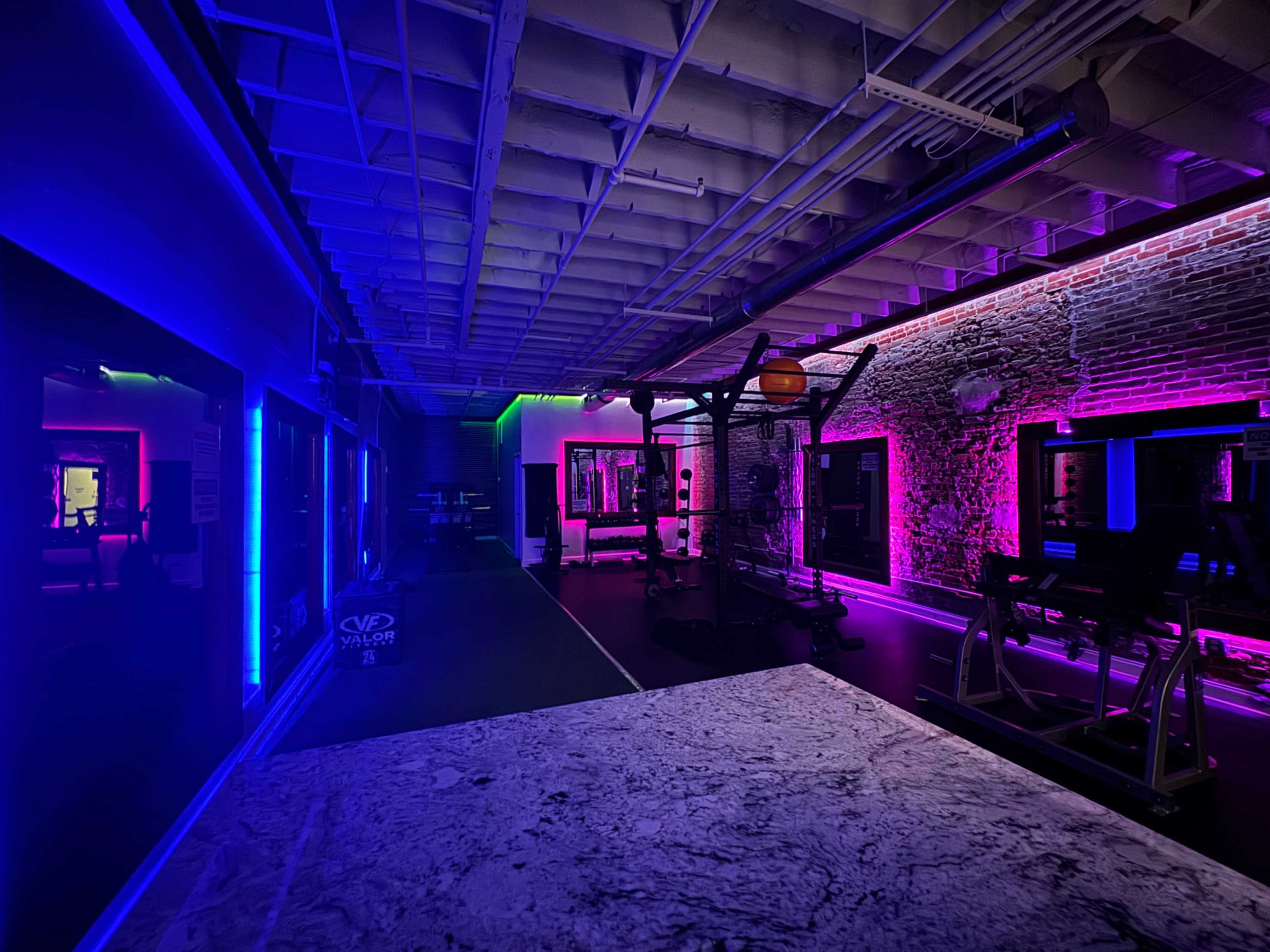 The image shows a gym interior lit with colorful neon lights in blue and pink, highlighting various workout equipment against a backdrop of exposed brick walls.