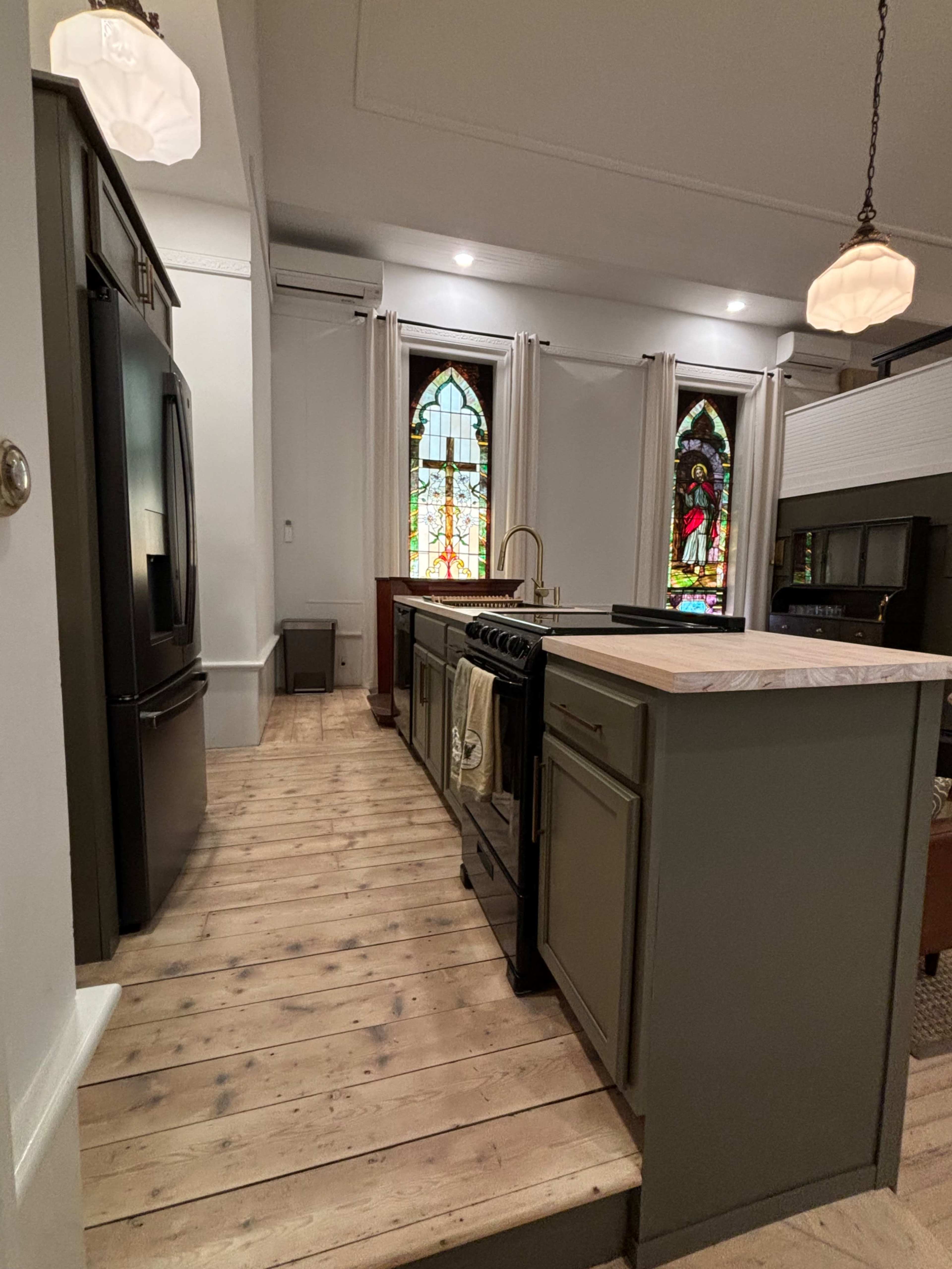 A kitchen features green cabinetry, a black stove, and a stained glass window, with wooden flooring and soft lighting.