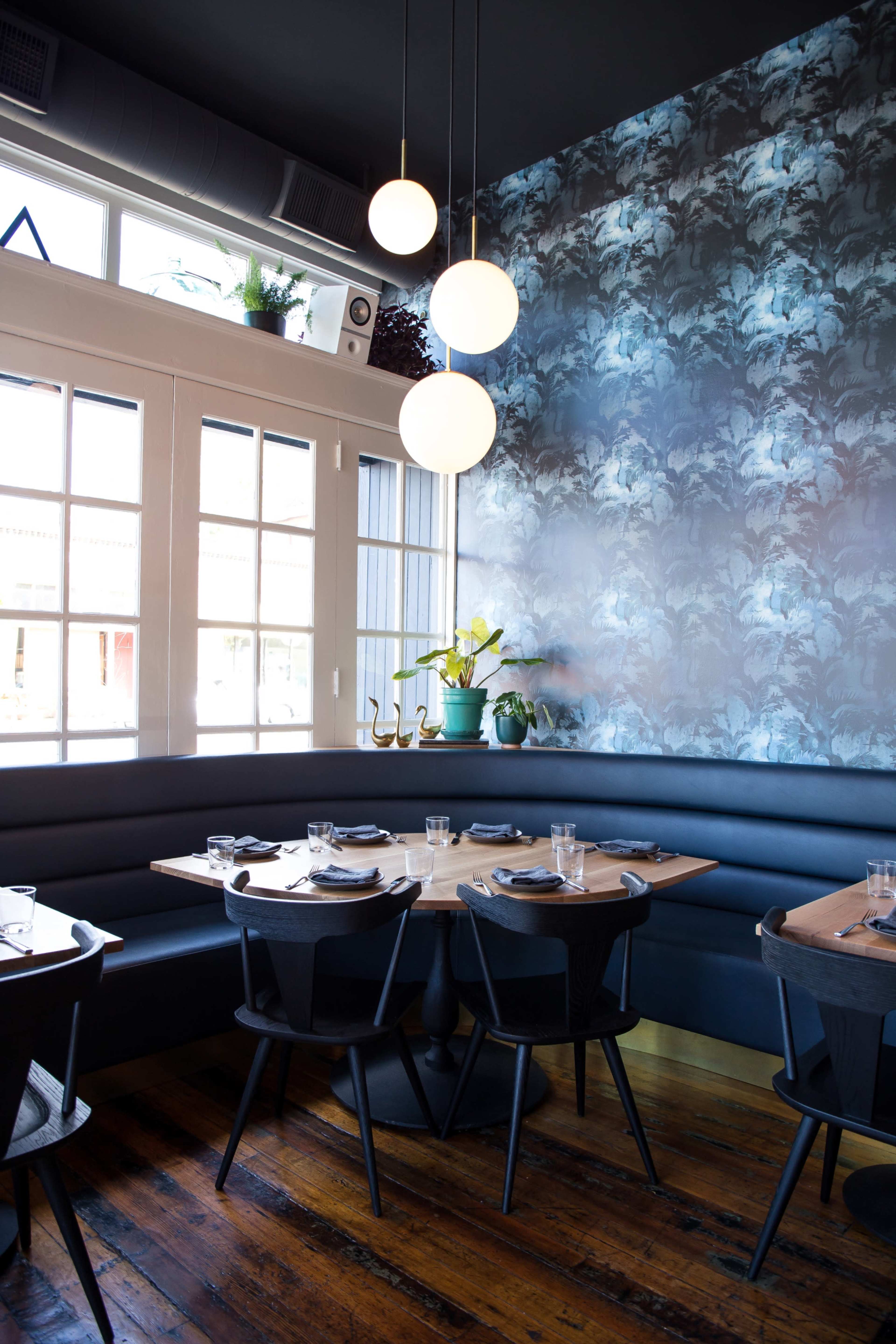 A modern restaurant interior featuring a dark wall with floral wallpaper, a booth seating area, and pendant lights hanging from the ceiling.