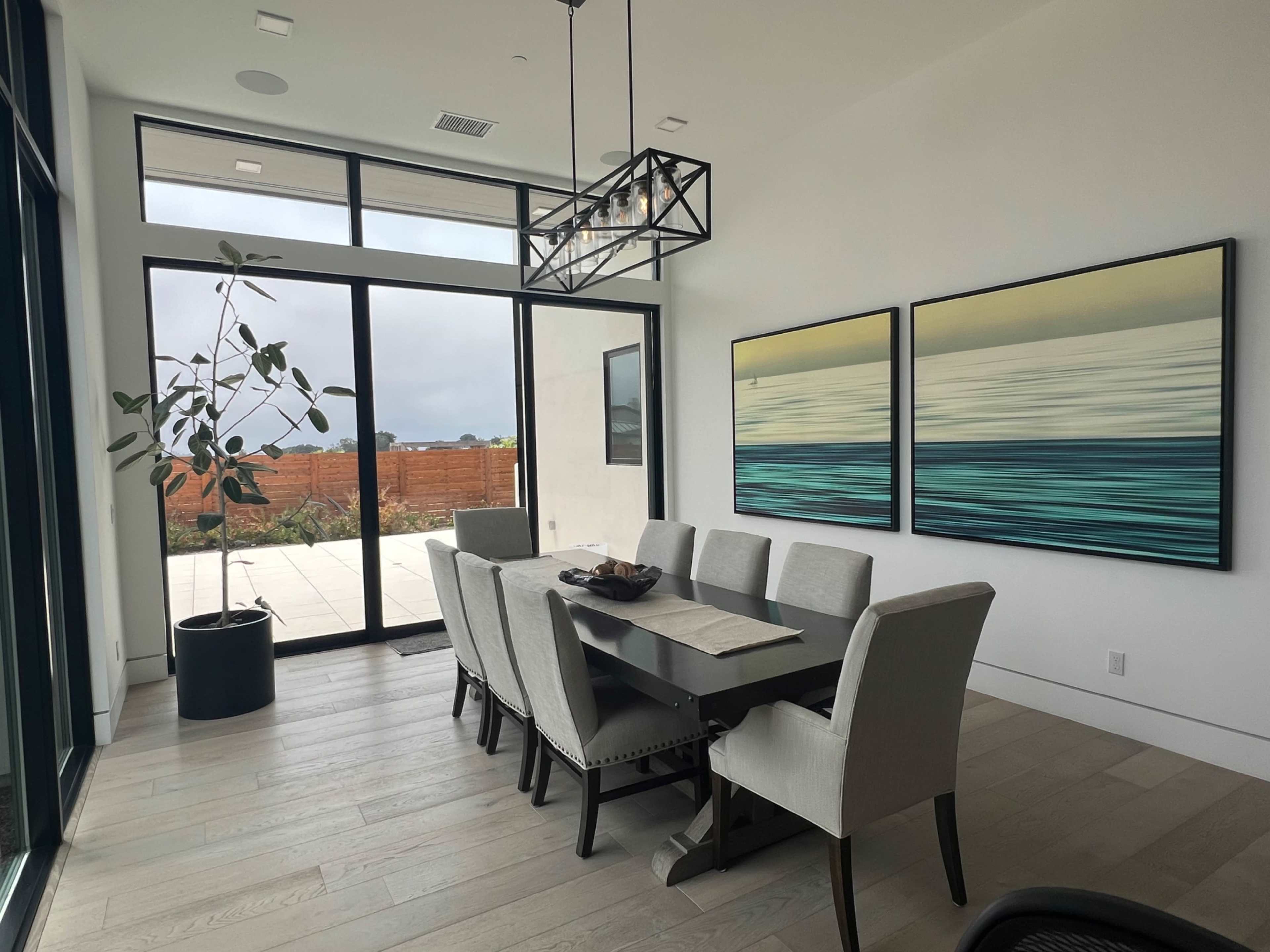 A modern dining room features a large table surrounded by upholstered chairs, with two abstract paintings on the wall and large windows providing a view of the outdoors.