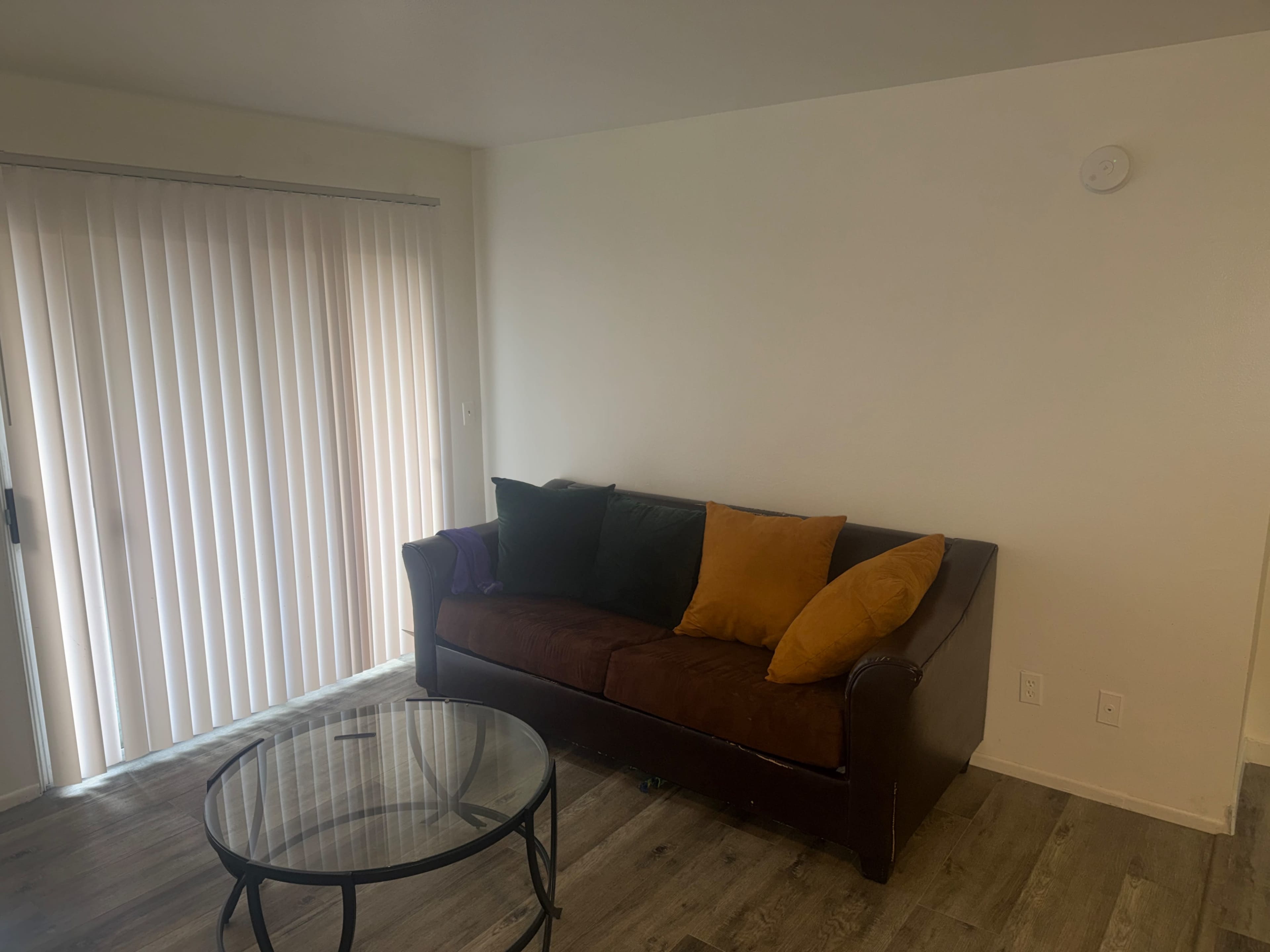 A brown sofa with colorful pillows is positioned in a room beside a set of vertical blinds and a glass coffee table.