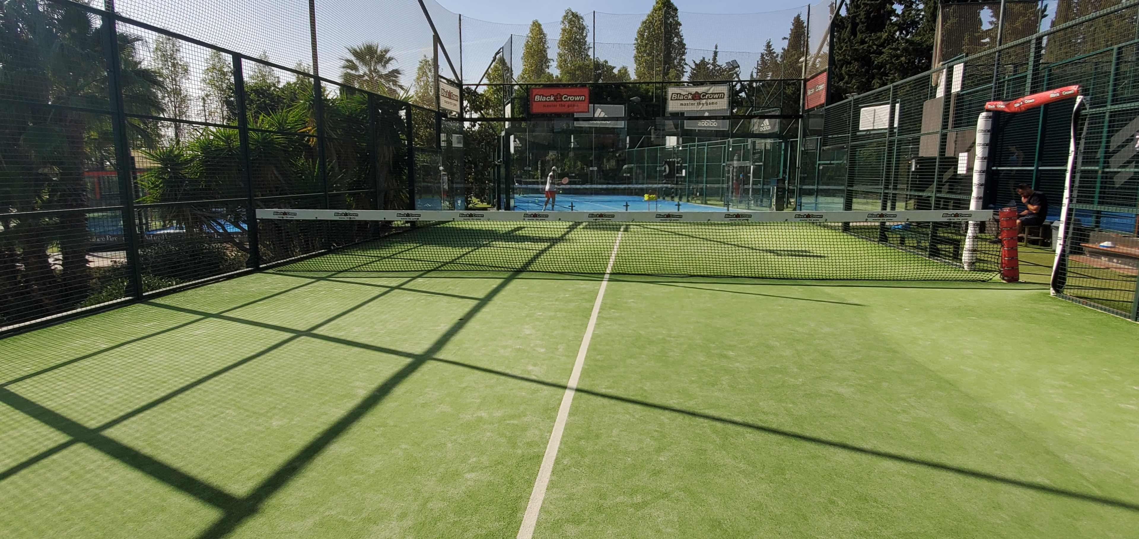 A well-maintained padel court with a green surface and surrounding netting, featuring players in the background.