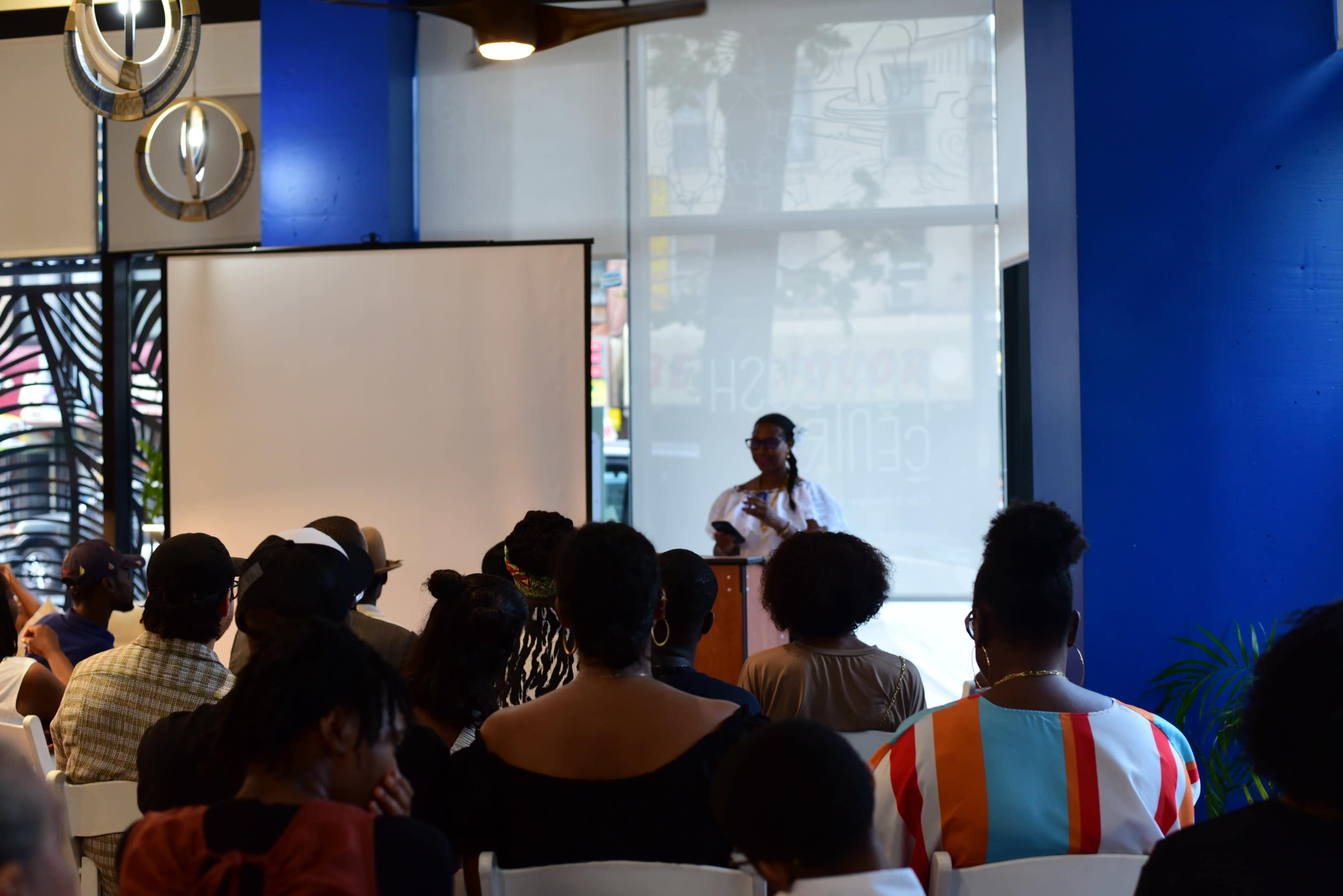A speaker addresses an audience in a brightly lit room with blue walls and a large screen in the background.