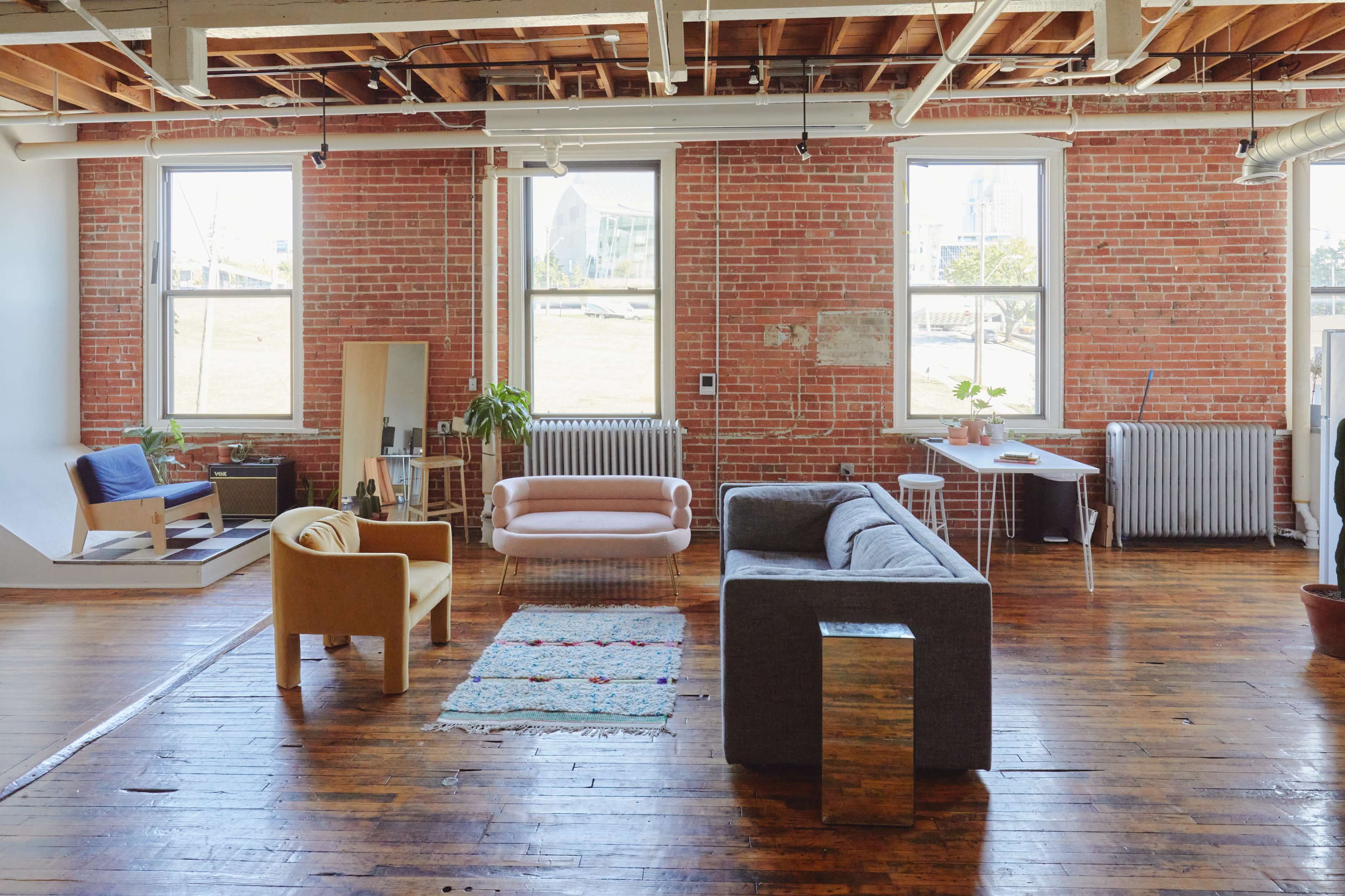 A spacious loft-style living area with exposed brick walls, hardwood floors, and various seating arrangements, including a gray sofa and a pink chair, along with plants and windows allowing natural light.